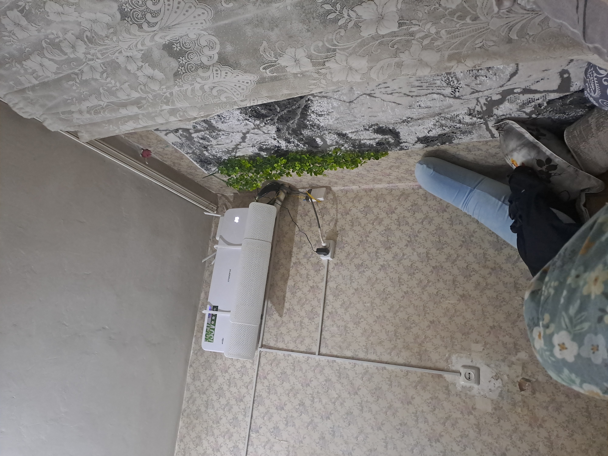 The image shows an indoor scene, likely a room, with a focus on a white air conditioner mounted on a wall with floral patterned wallpaper. A white electrical outlet is visible on the lower left of the wall, with peeling wallpaper around it. A black power cord runs from the air conditioner to a white adapter plugged into an outlet. A green vine with small leaves hangs down from the top of the wall near the center right. To the right of the vine, a portion of a wall tapestry with a black and white pattern is visible, partially covered by a white lace curtain. In the foreground, a person's legs, wearing light blue patterned pajama bottoms, are visible. Part of a blue bolster pillow, a dark garment, and other pillows are also present in the lower right of the frame, suggesting a casual or lived-in space. The scene depicts a domestic interior setting.