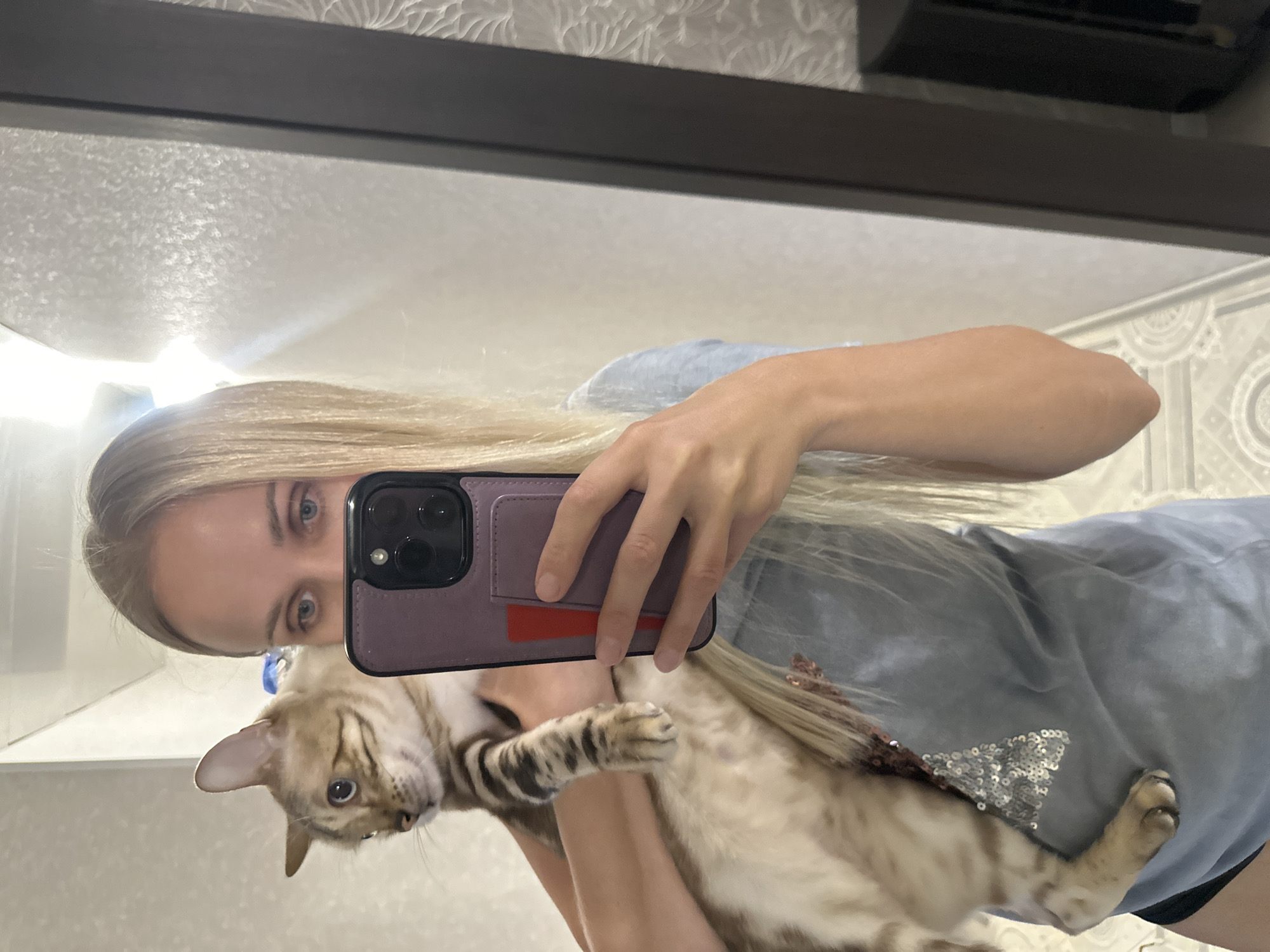 A woman with long blonde hair is holding a light-colored cat with dark stripes and blue eyes. The woman is taking a selfie in a mirror. She is wearing a light blue t-shirt with sequins on it. The cat is looking to the left. The background appears to be an indoor setting with a patterned wall and a dark framed mirror. The scene is not identifiable as being in Energetik, Russia.