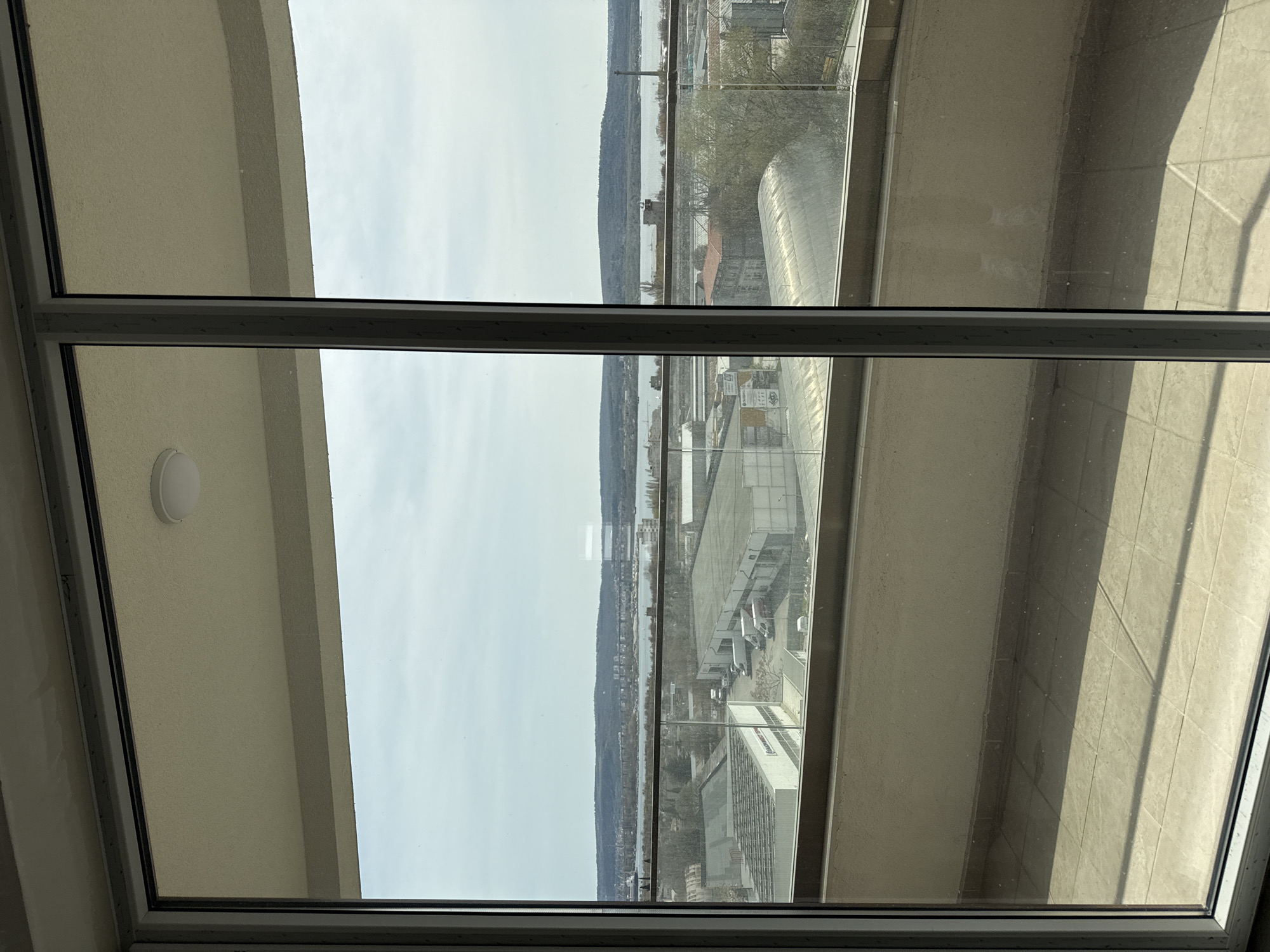 The image depicts a view from inside a building in Varna, Bulgaria, looking out through a large window. The window frame is made of gray metal. Outside, the cityscape includes buildings, roads, and a body of water with visible land on the opposite shore. There are several buildings with modern architecture, including one with a curved roof. The sky is overcast. Light from the sun is cast on the building interior. A light fixture can be seen on the wall inside the building.
