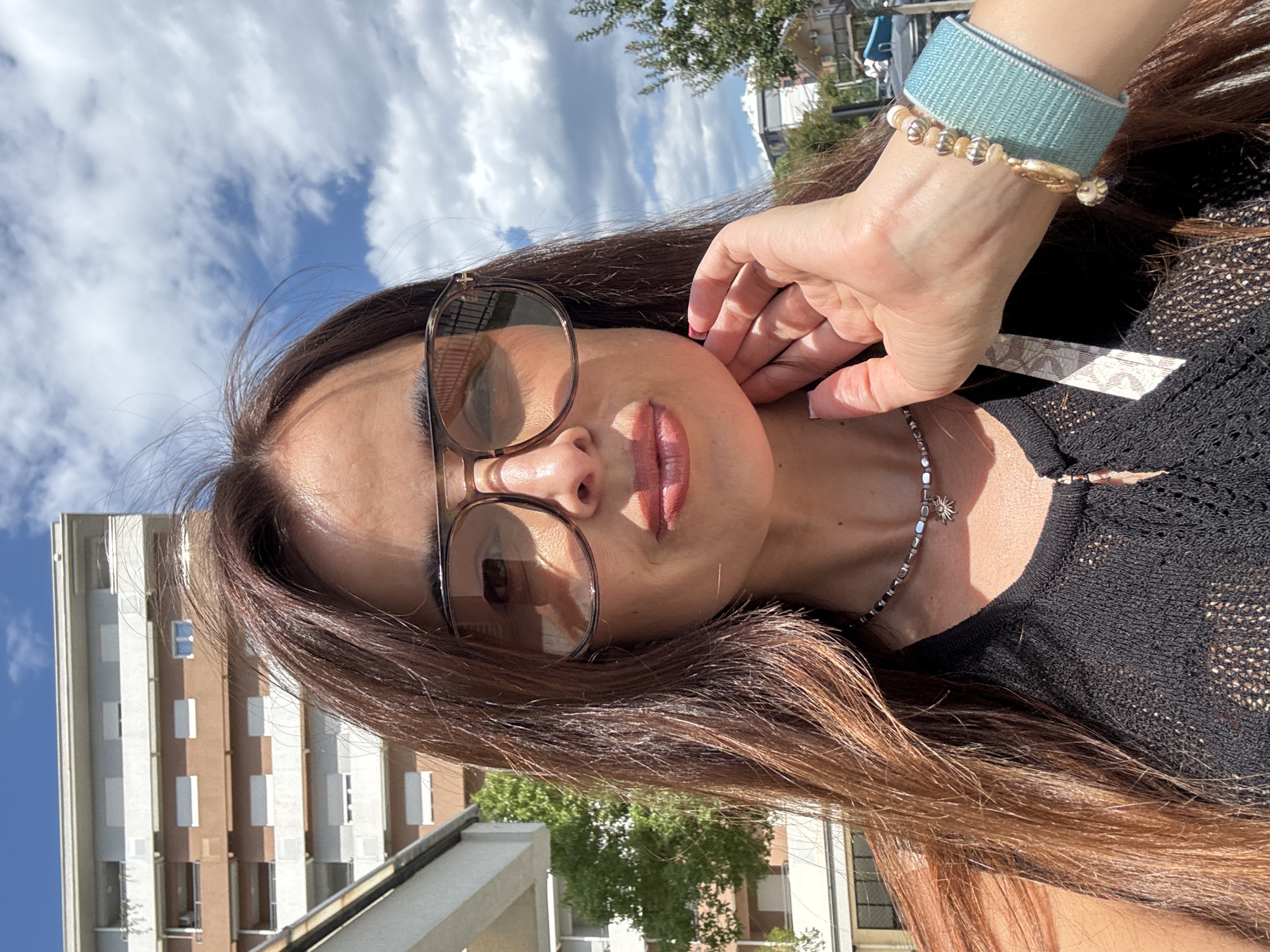 A medium shot captures a woman with long, brown hair, wearing oversized sunglasses and a black, sleeveless, open-knit top. She is outdoors, with a modern, multi-story building and trees in the background under a partly cloudy blue sky. Her right hand is raised to her chin, and she is wearing a silver necklace with a sun pendant and a light blue beaded bracelet. The woman's mouth is slightly open and her lips appear glossy.