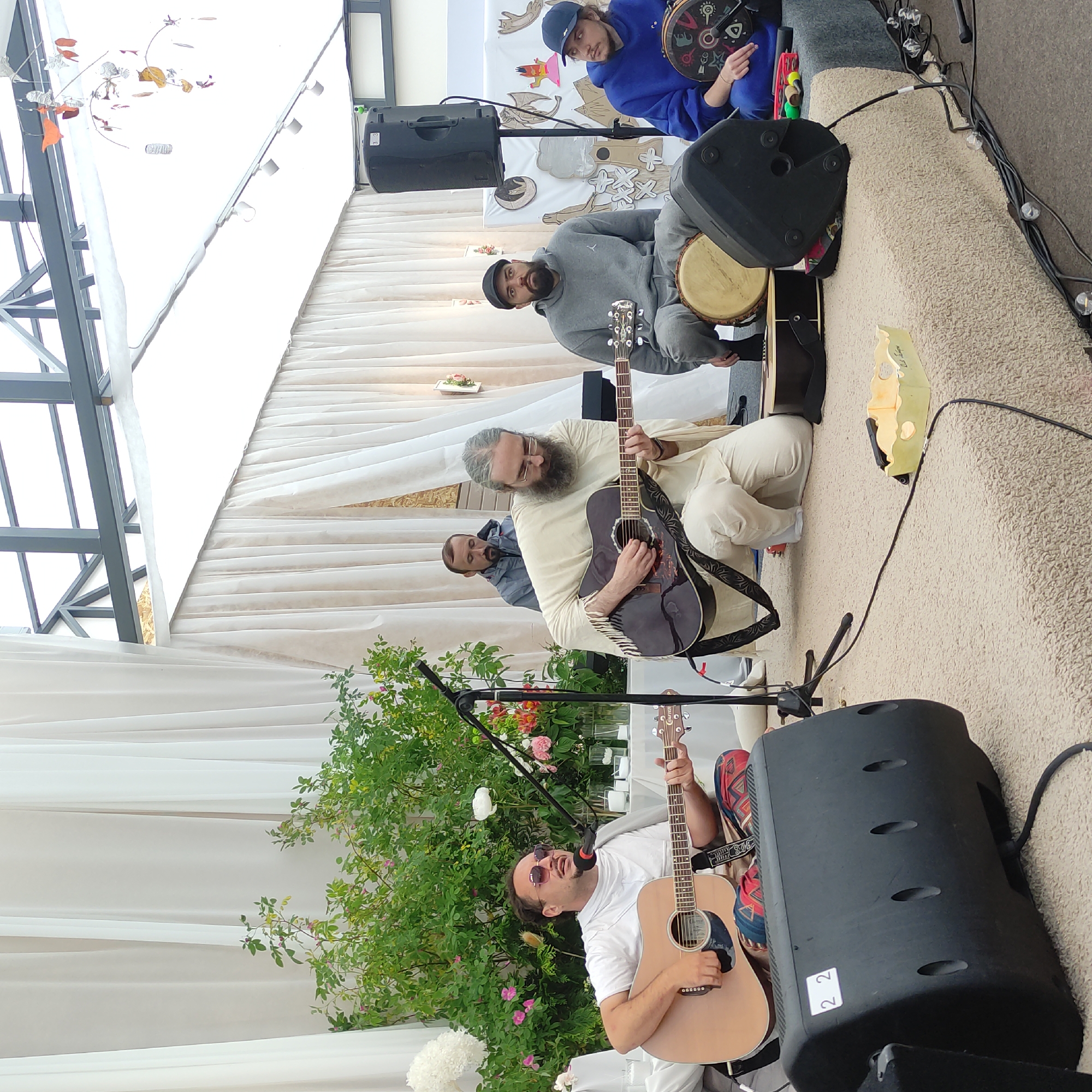 In Zhirnosovo, Russia, a group of five men are performing music outdoors. The scene appears to be a stage area, possibly at an event or festival, with white drapes forming a backdrop and a covered ceiling overhead.
In the foreground, a man with sunglasses and curly hair is playing an acoustic guitar and singing into a microphone. He is wearing a white t-shirt and colorful patterned pants. To his right, another man with a long beard and glasses is playing a dark acoustic guitar. He is dressed in a flowing cream-colored robe and pants. Behind him, a man in a gray hooded sweatshirt and pants is seated on a drum. To the far right, a man in a blue hooded sweatshirt and cap is holding a drum. Another man, partially obscured, stands behind the guitarist in the center.
Several musical instruments are visible, including two acoustic guitars and various drums and percussion instruments. Audio equipment, including speakers and microphones, is also present on the stage. The setting includes a lush green bush with pink and white flowers, suggesting an outdoor, possibly garden, environment.