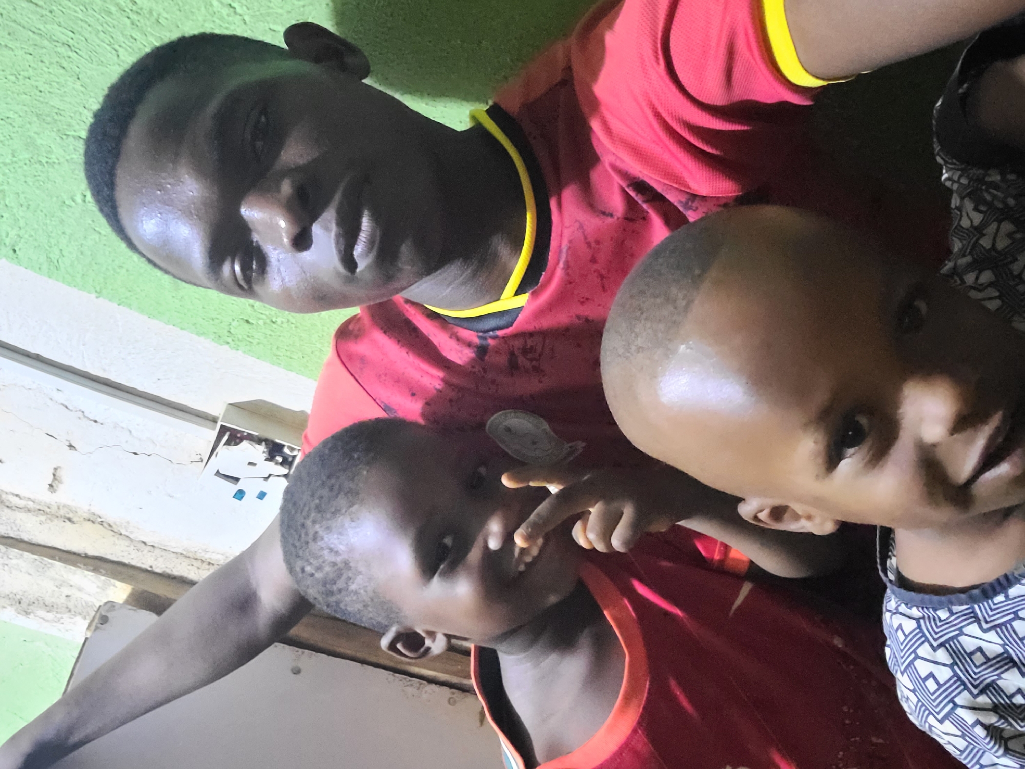 The image shows a man in a red shirt with yellow trim posing for a selfie with two children. The location appears to be indoors, with a green wall visible in the background. The lighting is somewhat dim. The man is looking at the camera with a neutral expression. One child is making a hand gesture, while the other is also looking at the camera. The angle of the shot suggests it was taken at close range.
