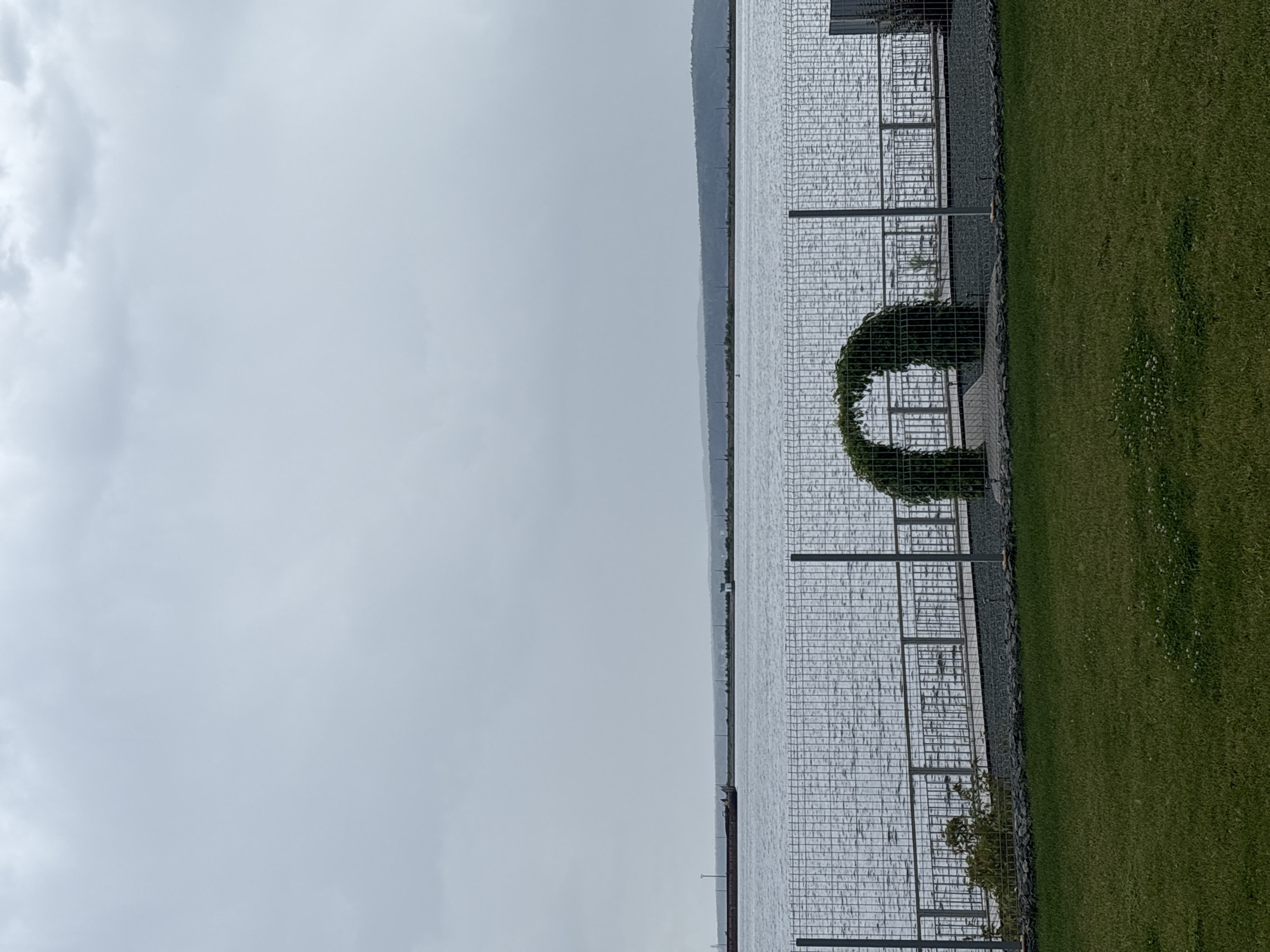 The image shows a cloudy sky over a body of water, likely a river or bay, with a distant shoreline. In the foreground, there is a grassy area and a metal fence. Beyond the fence, there is a walkway with a railing and an arched trellis covered in greenery. A barge is visible on the water in the distance. The location appears to be a waterfront area in Chaikovsky, Russia, possibly a park or public space.