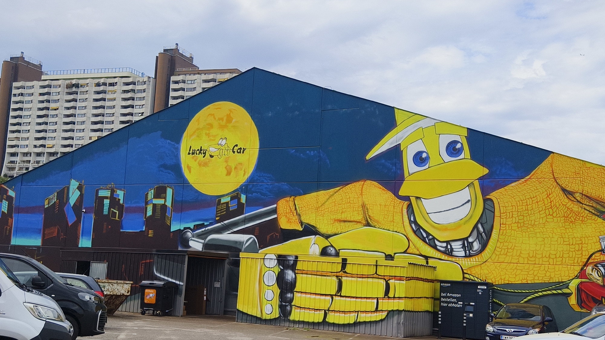 The image shows a large, colorful mural on the side of a building, depicting a cartoon character that appears to be a car. The character is predominantly yellow with large blue eyes and a wide smile. It is set against a dark blue night sky with a large yellow moon in the background, which has "Lucky Car" written on it. The mural also includes a stylized cityscape. In the foreground, there are several parked cars, a dumpster, and an Amazon Hub locker station. The building itself is a large industrial or commercial structure, and in the background, there are tall apartment buildings, characteristic of Vienna, Austria. The sky is overcast with scattered clouds.