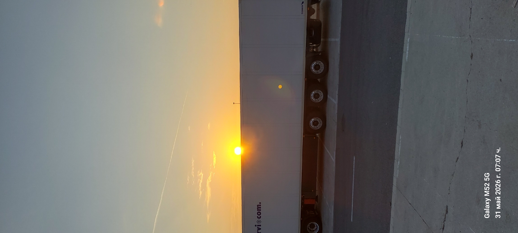 The image depicts a white trailer truck parked on a paved surface, likely a parking lot or loading area. The sun is setting or rising in the background, casting a bright orange glow that is partially obscured by the trailer. The truck's wheels and chassis are visible. To the right of the truck is a concrete or asphalt surface with cracks and seams. The text "Galaxy M52 5G 31 май 2026 г. 07:07 ч." is visible in the lower right corner, indicating the image was taken with a Galaxy M52 5G phone on May 31, 2026, at 07:07. The location is provided as Alzira, Spain.