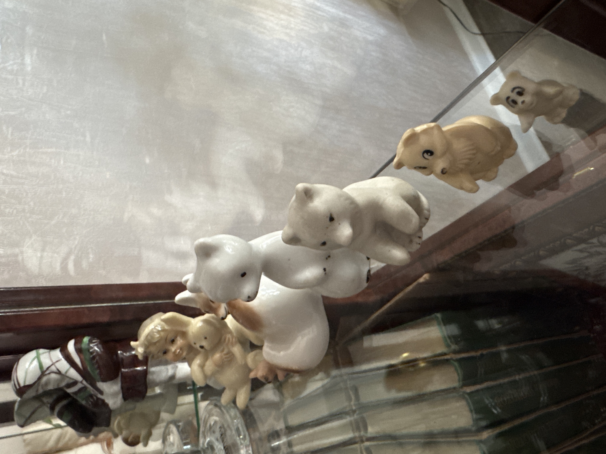 The image depicts a collection of ceramic figurines displayed on a glass shelf within a wooden cabinet. The figurines include several teddy bear-like figures in various sizes and colors, a young child holding a bear, and a white bird-like figurine. The background is a white, textured surface, possibly a curtain or wall covering. To the left, a glimpse of books with dark green spines is visible. The lighting suggests an indoor setting. There are no people present in the scene. The location is identified as Novosibirsk, Russia.
