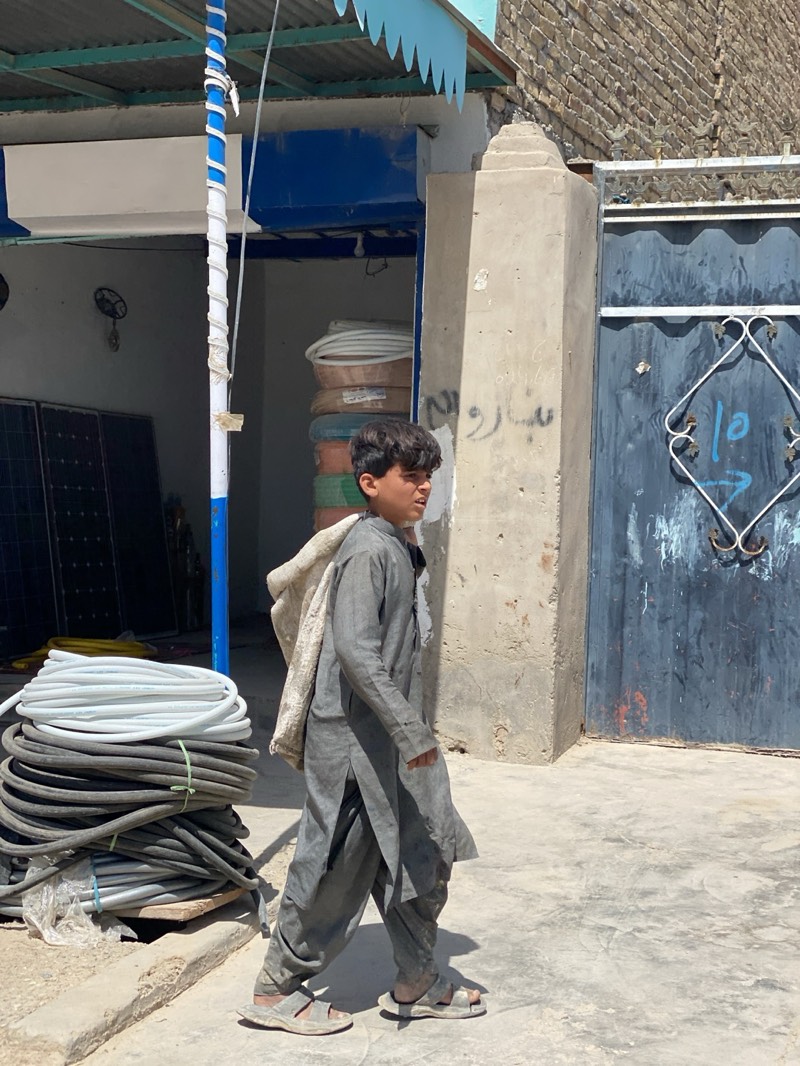 A boy is walking on a sidewalk, carrying a bundle over his shoulder in the city of Kandahar, Afghanistan. The boy is dressed in a dirty grey shalwar kameez and sandals. He is walking to the right, away from a building with exposed electrical and solar equipment. In the background, is a gate with Arabic writing on the post.