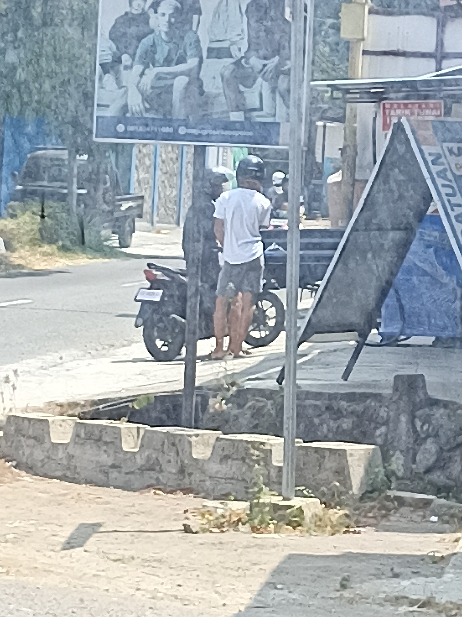 The image depicts an outdoor scene, possibly in the city of Pulisen, Indonesia, characterized by a street, sidewalk, and some vegetation. In the foreground, a low, weathered stone wall with carved recesses runs horizontally. Beyond this, a paved area transitions to a road where a black motorcycle is parked. To the right of the motorcycle, a person wearing a white t-shirt and dark shorts stands, facing away from the camera. Behind this individual, a dark silhouette of another person is visible.
In the background, a large billboard or poster with images of several people is displayed, partially obscured by trees and a blue tarp. Several other signs are also present, one of which appears to be a red and white sign with text. A light-colored pole stands prominently in the mid-ground, near the person in white. The overall lighting suggests a daytime scene with bright sunlight casting shadows. The environment appears somewhat arid, with dry grass and sparse vegetation visible.