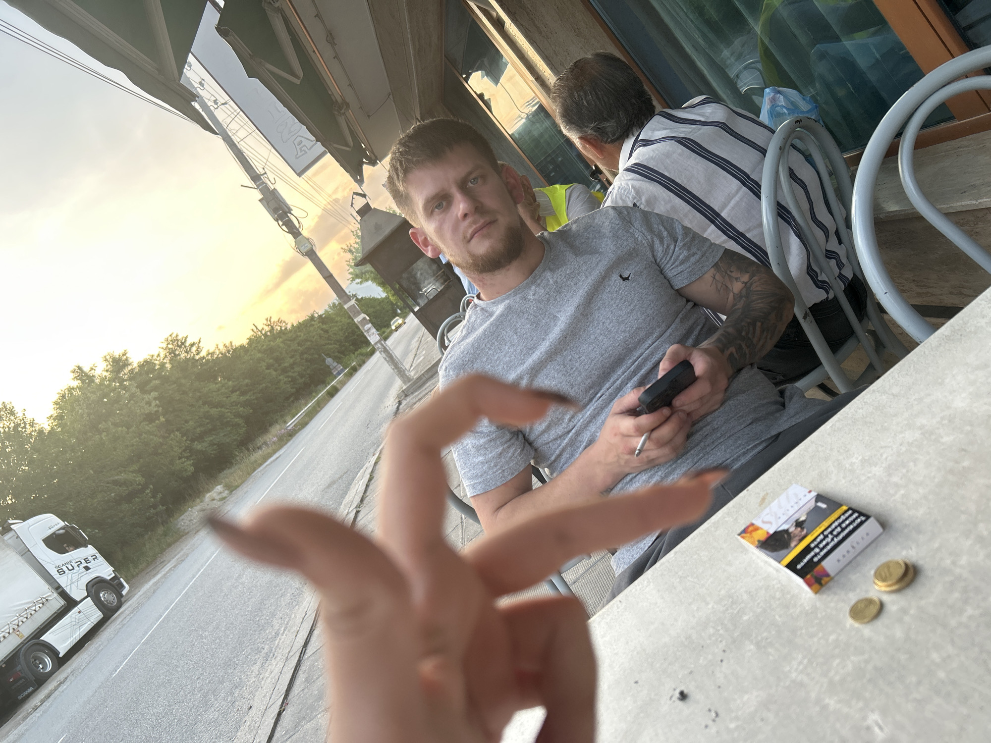 The image depicts an outdoor scene at golden hour, likely a cafe or restaurant patio situated alongside a road in the city of Unknown, Unknown. In the immediate foreground, a blurry hand with dark nail polish extends towards the viewer. On a light grey concrete or stone table, a pack of Karelia cigarettes with a warning label and two stacked gold-colored coins are visible.
In the midground, a man with short brown hair, a beard, and light eyes looks directly at the camera. He wears a grey t-shirt with a small dark emblem on the chest and has a tattooed sleeve on his left arm. He is seated at the table, holding a black smartphone and a lit cigarette in his right hand. Behind him, a person partially obscured wears a yellow garment. To the right of the central man, another person is seated with their back mostly to the camera, wearing a white and dark-striped short-sleeved shirt. The chairs are metal with light grey, rounded frames.
The background shows an asphalt road with white markings, where a white Scania semi-truck with "SUPER" written on its side is visible, facing away from the camera. Beyond the road, green trees and foliage line the area. The sky above displays vibrant sunset colors, with bright yellow and orange hues near the horizon. To the right, part of a building with large reflective windows and a green awning is seen, along with a utility pole and a dark, possibly industrial or grill-like structure. Reflections of the sky and surroundings are visible in the building's windows.