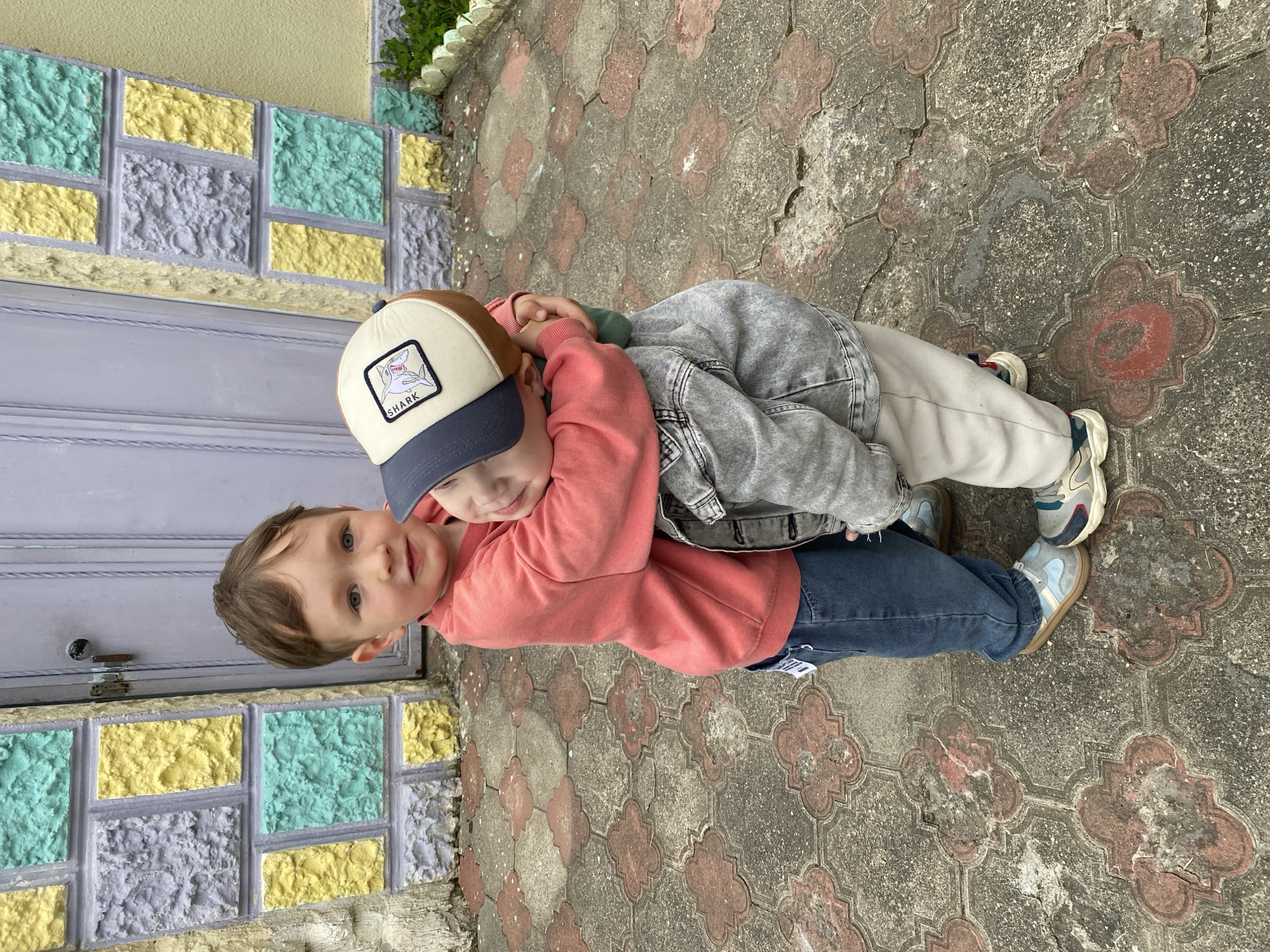 Two young children are embracing. One child wears a salmon-colored long-sleeve shirt, blue jeans, and a baseball cap with a shark logo. The other child wears a grey denim jacket and light-colored pants. They are standing on a paved area with interlocking patterned stones. In the background is a wall made of textured, colorful blocks (yellow, teal, purple). The scene appears to be outdoors, possibly in a residential area of Sochi, Russia.