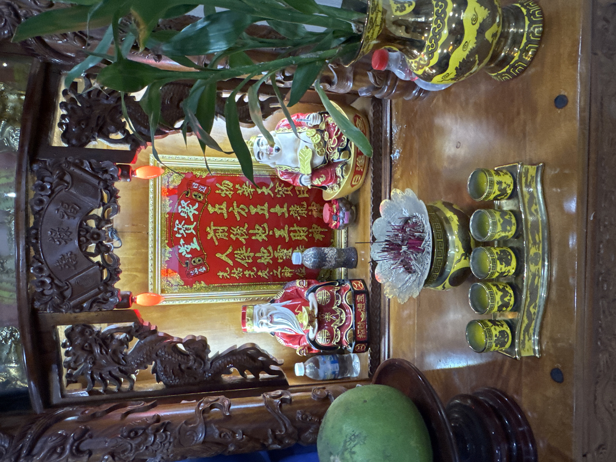 The image depicts an altar within a shrine, likely a Buddhist or Taoist temple, in Nha Trang, Vietnam. The altar is made of dark, polished wood and features intricate carvings, including a dragon on the left side and decorative scrollwork on the top. At the center of the altar, a red scroll with gold Chinese characters is displayed. Flanking the scroll are two statues of deities, likely representing prosperity or good fortune. The statue on the left is seated, wearing red robes and holding a gold ingot. The statue on the right is also seated, dressed in similar red attire and holding a fan.
In front of the statues, an incense burner with burning incense sticks is placed on a decorative golden stand. To the left of the incense burner, a green pomelo rests on a wooden plate, and further left, two plastic bottles of water are visible. To the right of the incense burner, a set of four small, golden cups with dragon motifs are arranged on a tray. A tall, ornate vase with a yellow and black pattern, containing a plant with green leaves, stands on the right side of the altar. Several plastic bottles are also present on the altar. The lighting appears to be from overhead sources and red decorative lights, creating a warm, ambient glow. The overall scene suggests a place of worship or veneration, common in religious sites across Vietnam.