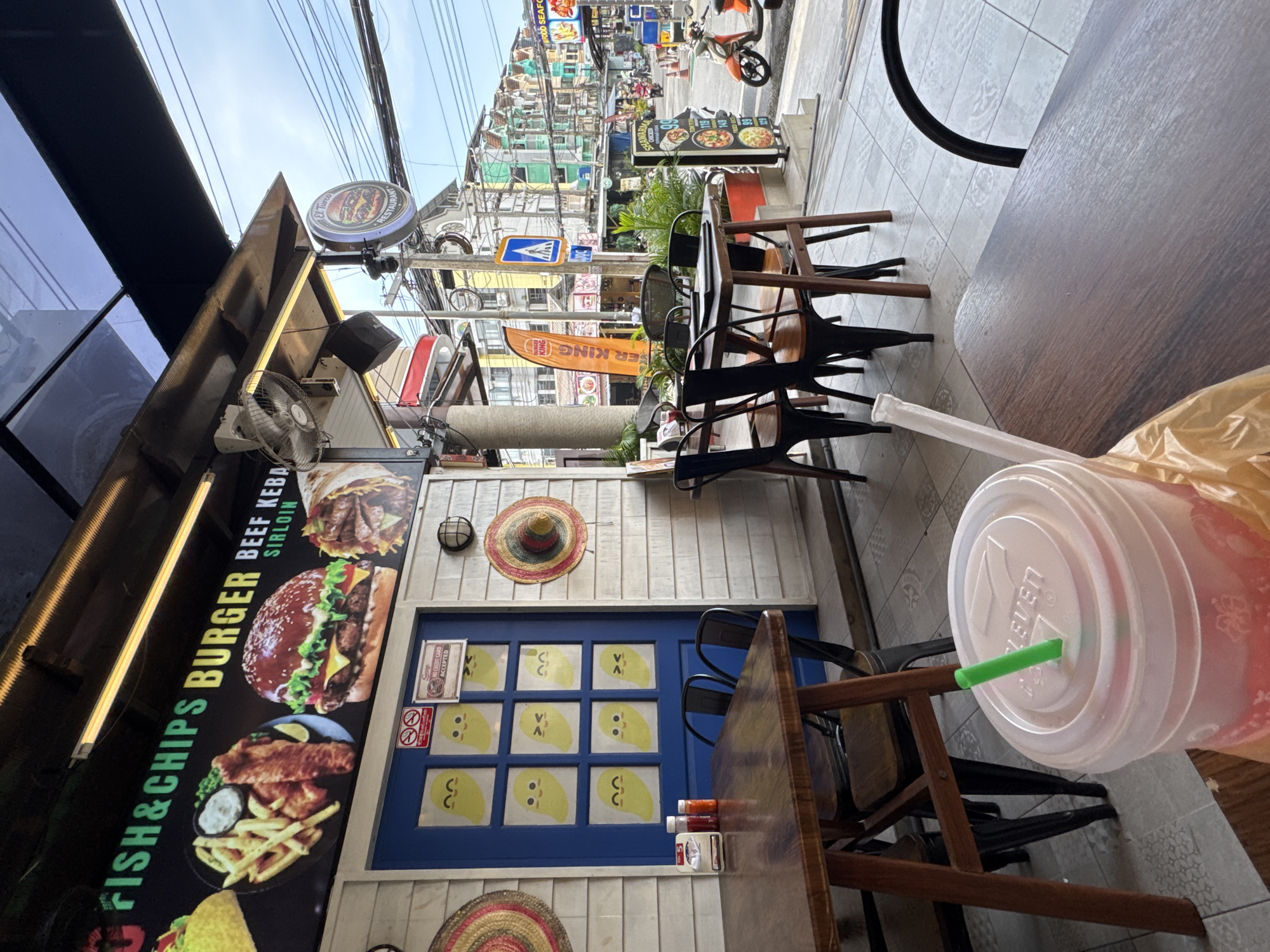 A street-level exterior shot shows a cafe with tables and chairs arranged outside. In the foreground, a large plastic cup with a green straw sits on a wooden table. Behind it, another wooden table with four black chairs is visible. The cafe's exterior wall is white with a blue door adorned with cartoon mango characters. A sign above the cafe reads "FISH & CHIPS BURGER BEEF KEBAB SIRLOIN." Further down the street, colorful buildings, power lines, and various signs indicate a commercial area. An orange scooter is parked on the right side of the street. The scene appears to be located in Karon, Thailand, given the architecture and signage.