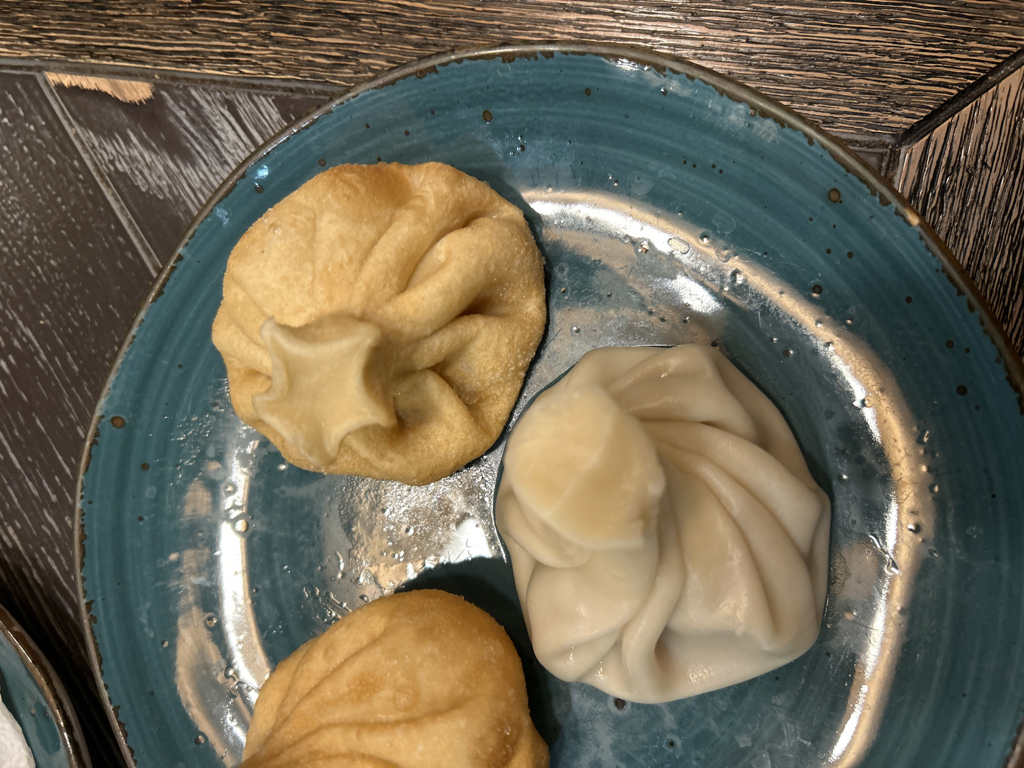 The image shows a close-up, high-angle shot of three dumplings arranged on a blue speckled plate. The plate is placed on a wooden surface with a rustic finish. Two of the dumplings are golden brown and appear to be fried, while the third dumpling is paler in color, possibly steamed or boiled. The dumplings have a pinched or folded appearance at the top. There are small droplets of liquid on the plate, possibly water or oil. The photograph appears to be taken indoors with natural or artificial light. No people are visible in the image. There is no information in the image connecting it to Izhevsk, Russia.