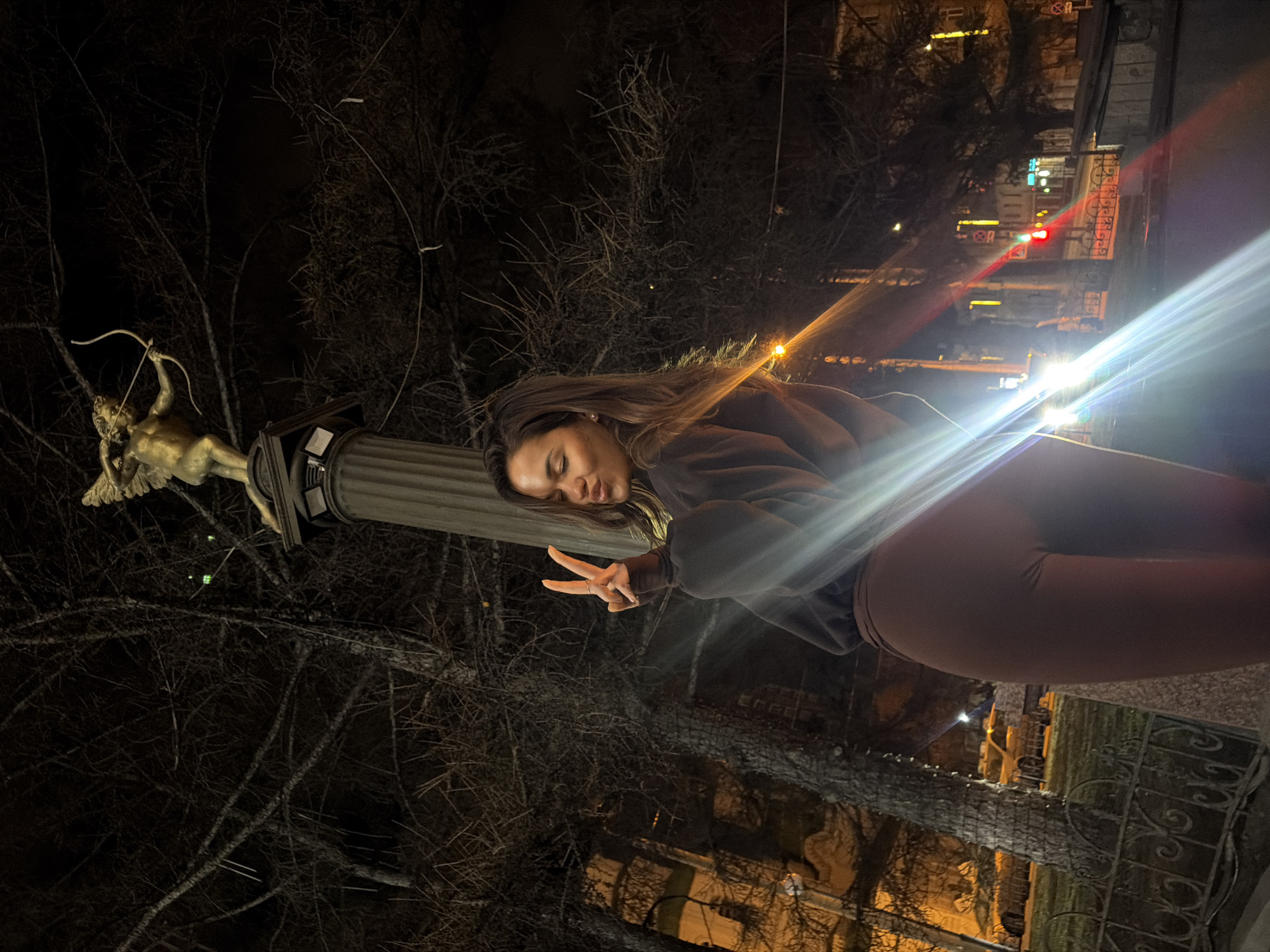 An outdoor, nighttime scene captures a young woman posing next to a statue and column, illuminated by artificial light sources. The image is taken at night, with the sky completely dark and various distant lights creating prominent lens flares across the frame.
The main subject is a young woman positioned on the right, facing towards the viewer. She has long, dark hair, wears a dark, long-sleeved top, and brown leggings. She is making a peace sign with her right hand, her eyes are closed or looking downwards, and she has a subtle, relaxed smile.
To her left stands a golden or bronze-colored statue atop a dark, fluted column. The statue depicts a cherubic figure, resembling Cupid, holding a bow and arrow as if poised to shoot.
The background consists of numerous bare tree branches, suggesting a winter or early spring setting. Beyond the trees, distant city lights from buildings and street lamps are visible, indicating an urban environment. A decorative metal fence or railing is present in the lower right and mid-ground. Faintly visible in the far background, among the city lights, is a red traffic light and a red circular road sign with a white horizontal bar, typically denoting "No Entry" or "Do Not Enter." The overall mood is casual and slightly playful.