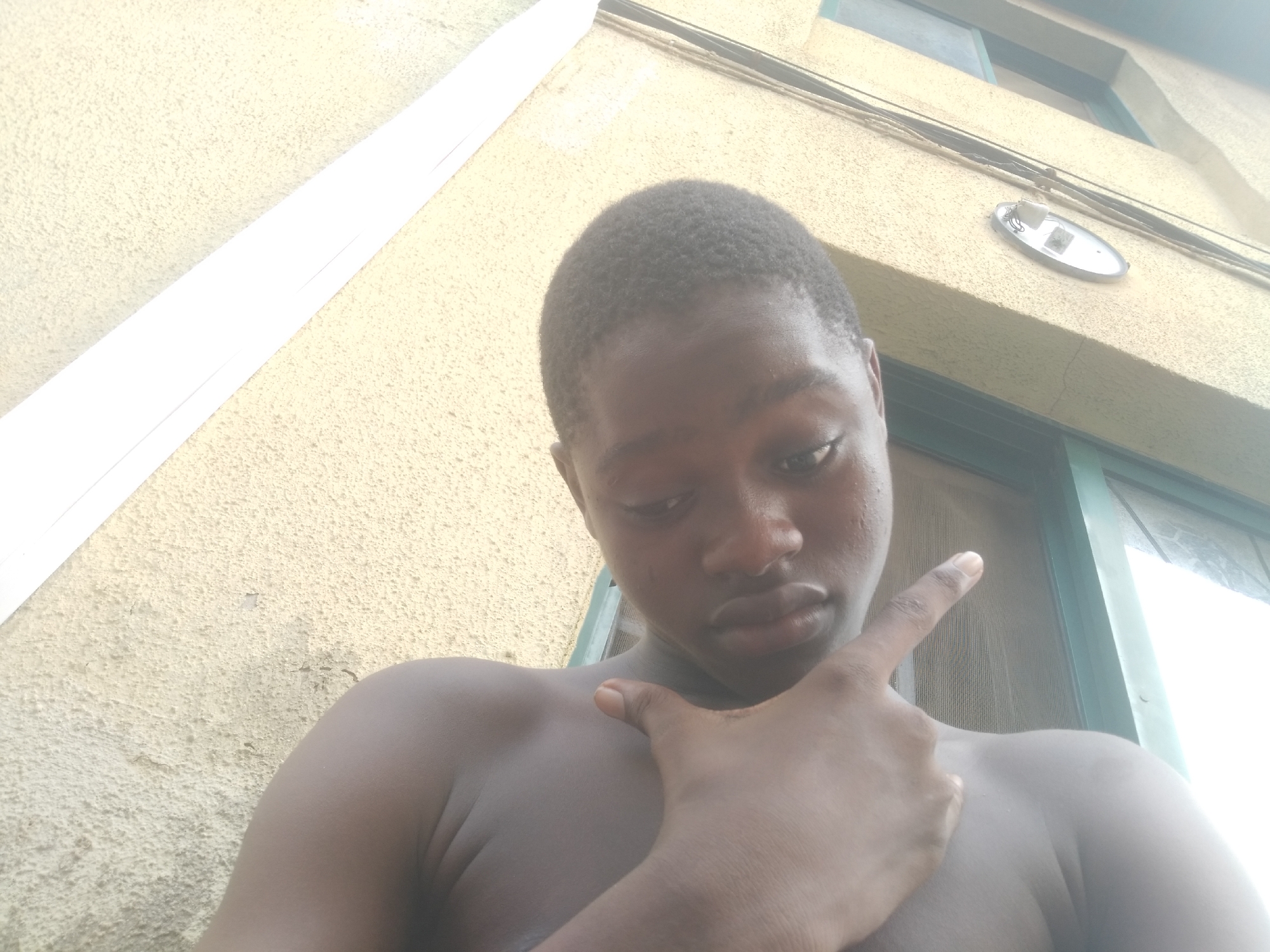 A shirtless dark-skinned young man is in the foreground of the shot, with his right arm across his chest. His index finger is extended upwards. The background shows the exterior of a yellow building with an off-white trim. There are some cables running along the wall and an oval-shaped light fixture. A window is partially visible in the upper right corner. No further information is available to verify a specific location.