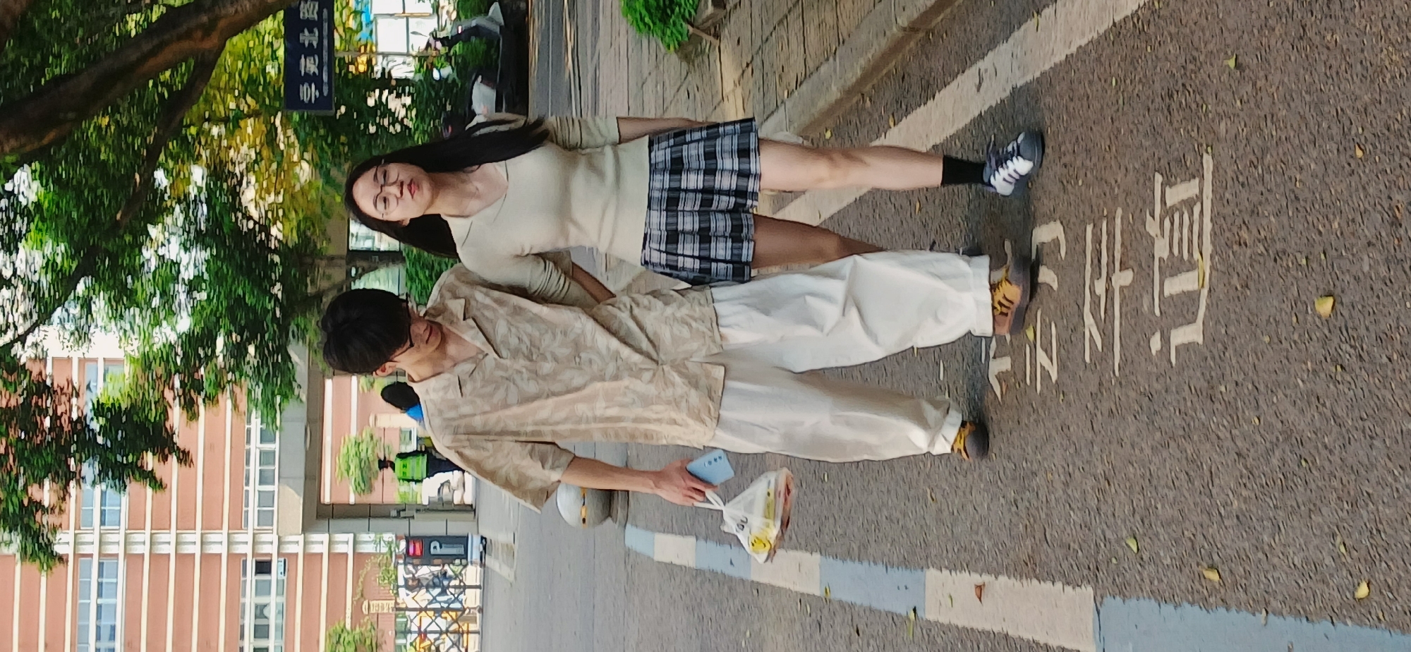 Two individuals, a male and a female, are standing on a crosswalk in Chongqing High-tech Zone, China. The female is wearing a light-colored long-sleeved top, a plaid skirt, black socks, and grey sneakers. The male is wearing a patterned short-sleeved shirt, white pants, and tan sneakers. He is holding a smartphone in his left hand and a plastic bag with some items in his right hand. The crosswalk has Chinese characters painted on the asphalt. The background includes trees, a street sign, and a building with multiple windows.