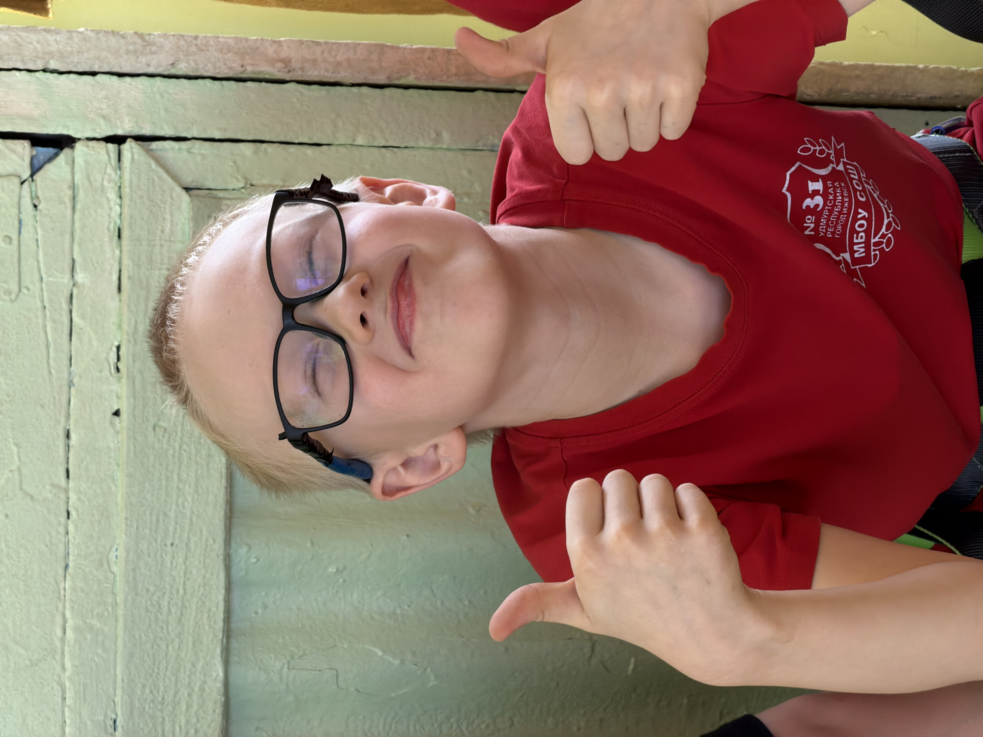 A young boy with blond hair and glasses stands in front of a pale green wooden structure. He wears a red shirt with a logo that includes the number "31" and the text "Удмуртская Республика Город Ижевск МБОУ СОШ" (Udmurt Republic, City of Izhevsk, MBOU SOSH). The boy gives a thumbs-up with both hands and closes his eyes while smiling. He has a blue object attached to his glasses near his left ear.