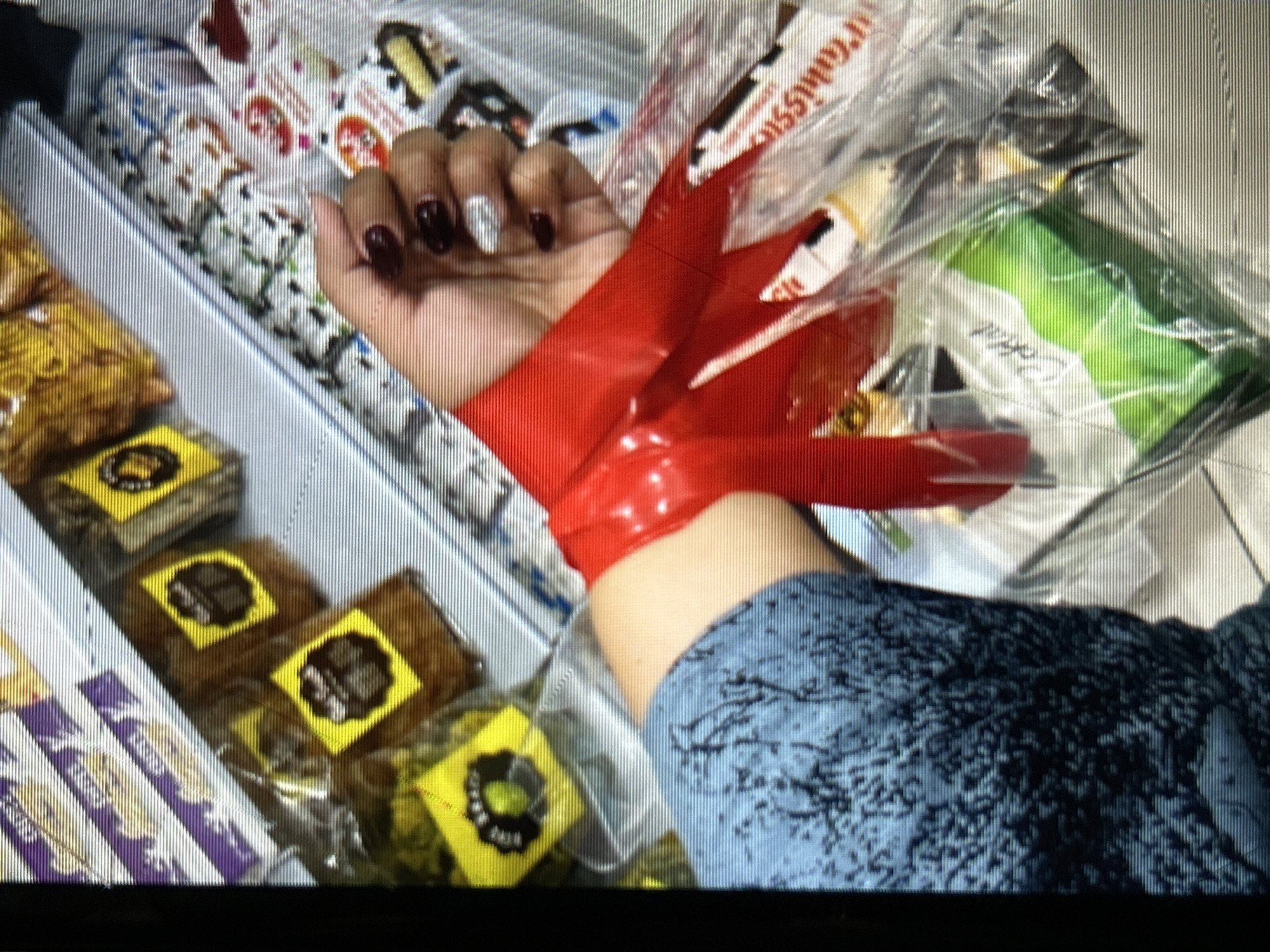 The image shows a person's arm, adorned with long, red, pointed, artificial fingernails, wrapped in red tape. The arm is positioned in front of a display of various packaged snacks and sweets. These items appear to be arranged in rows on shelves within a retail environment, possibly a convenience store or a market stall. Some of the packaging is transparent, revealing the contents, while others have colorful designs and labels.
The visible products include what look like individually wrapped cakes or brownies with yellow labels featuring a black circular logo. There are also other items with white and colorful packaging, some with text that is partially legible. In the background, there are more packages, some in clear plastic, one with a green label. The overall impression is a close-up shot focusing on the hand and arm, with the merchandise serving as a backdrop.
The lighting suggests an indoor setting, and there are no clear indicators of the time of day or weather conditions. The image seems to capture a specific, perhaps unusual, pose or an element of personal adornment in a commercial setting. The focus on the artificial nails and the red tape suggests a deliberate styling choice or a prop for an activity. No specific emotions are directly conveyed by the image, other than a potential sense of curated presentation.
There is some visible text on the packaging, though it is largely indistinct due to the image resolution and the angle. One prominent piece of text on a white wrapper appears to be "Tarbiss". Other labels are too blurry to read with certainty. The location context of Depok, Indonesia, does not provide specific visual cues within the image itself.
