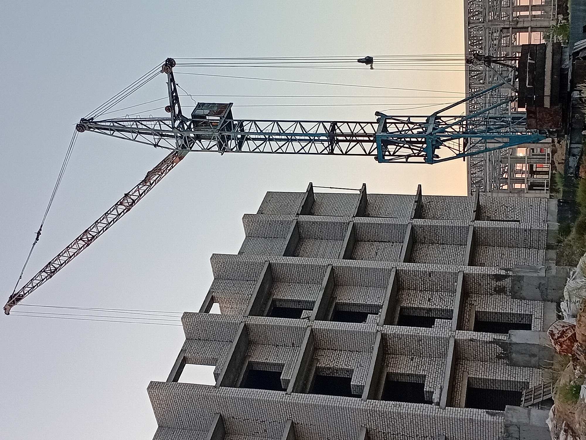 The image depicts a construction site in the city of Semey, Kazakhstan. A large tower crane, painted blue and red, dominates the scene, its boom extended over an unfinished brick building. The building, constructed with white bricks, has multiple stories with open window frames and concrete slabs. The crane's cab is visible at the top of its tower. In the background, another partially constructed structure with a metal framework can be seen. The sky is clear and pale, suggesting it may be early morning or late evening. The ground in the foreground is covered with soil, some debris, and what appears to be construction materials. There are no people visible in the image.