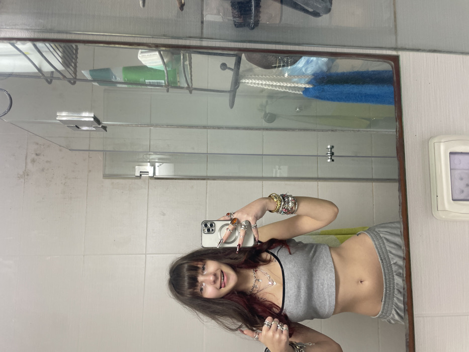 A young woman with bangs and dark hair with red highlights poses in a bathroom mirror selfie. She wears a grey crop top and grey sweatpants. Multiple rings adorn her fingers, and bracelets cover her wrists. A silver necklace rests on her collarbone. The bathroom features white tiled walls, a glass shelf with toiletries, and a large mirror with a dark brown frame. A white electrical outlet is visible on the right.