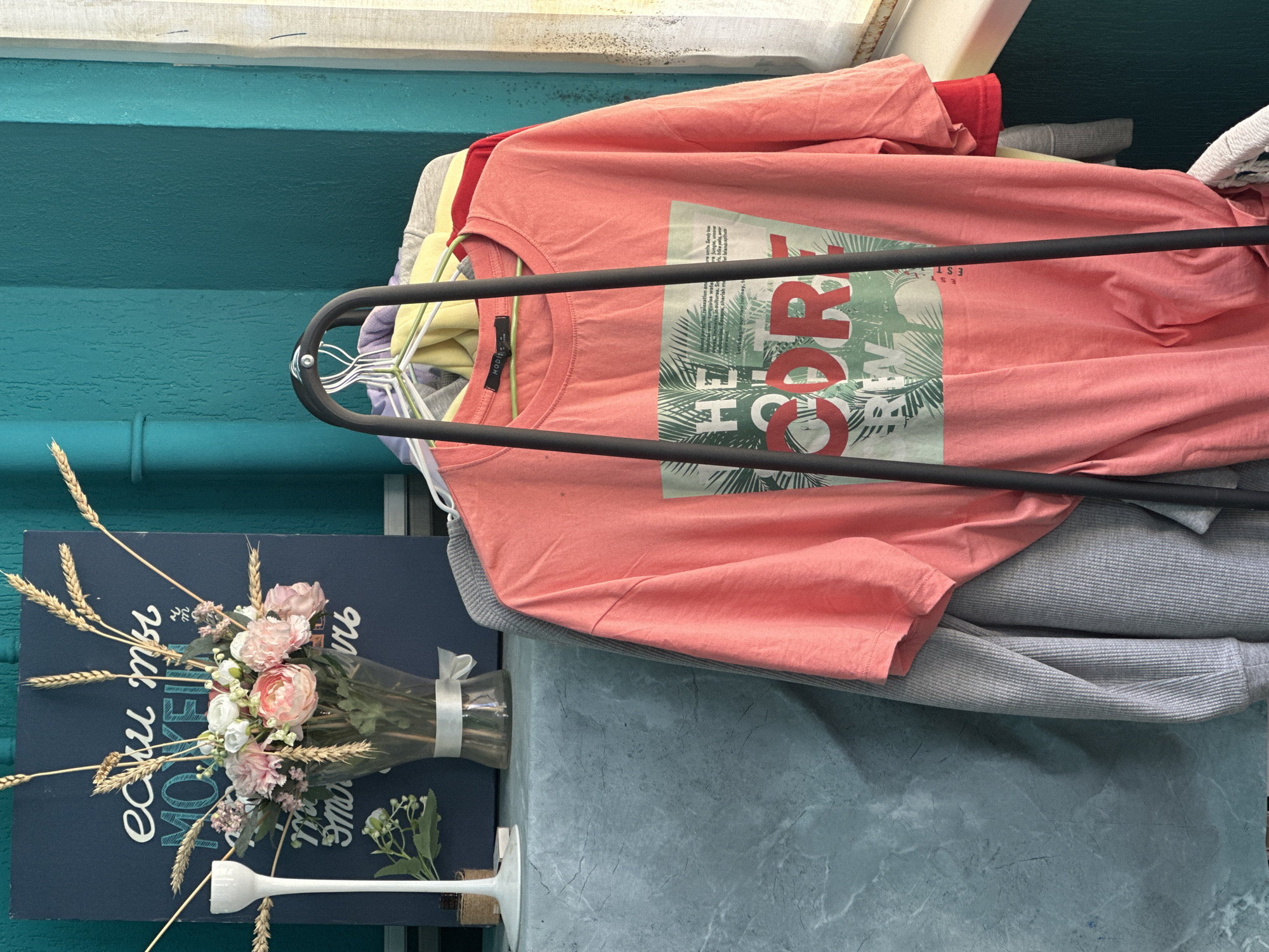 A close-up, eye-level shot captures a rack of clothing against a teal-colored wall. In the foreground, a vibrant coral t-shirt with a graphic print of palm leaves and red lettering hangs prominently. To the left of the t-shirt, a gray waffle-knit sweater is partially visible. Behind these, other garments in yellow, red, and purple are draped over hangers on the same rack. To the far left, a narrow white vase with a white ribbon and a bouquet of pink and white flowers, along with dried wheat stalks, sits on a textured gray surface. Behind the flowers, a dark blue sign with white lettering is visible. The scene appears to be indoors, possibly in a domestic setting or a small boutique in Sochi, Russia, suggested by the overall aesthetic and color palette, though no definitive Sochi landmarks are present. Natural light streams in from the right side of the frame, illuminating the scene.