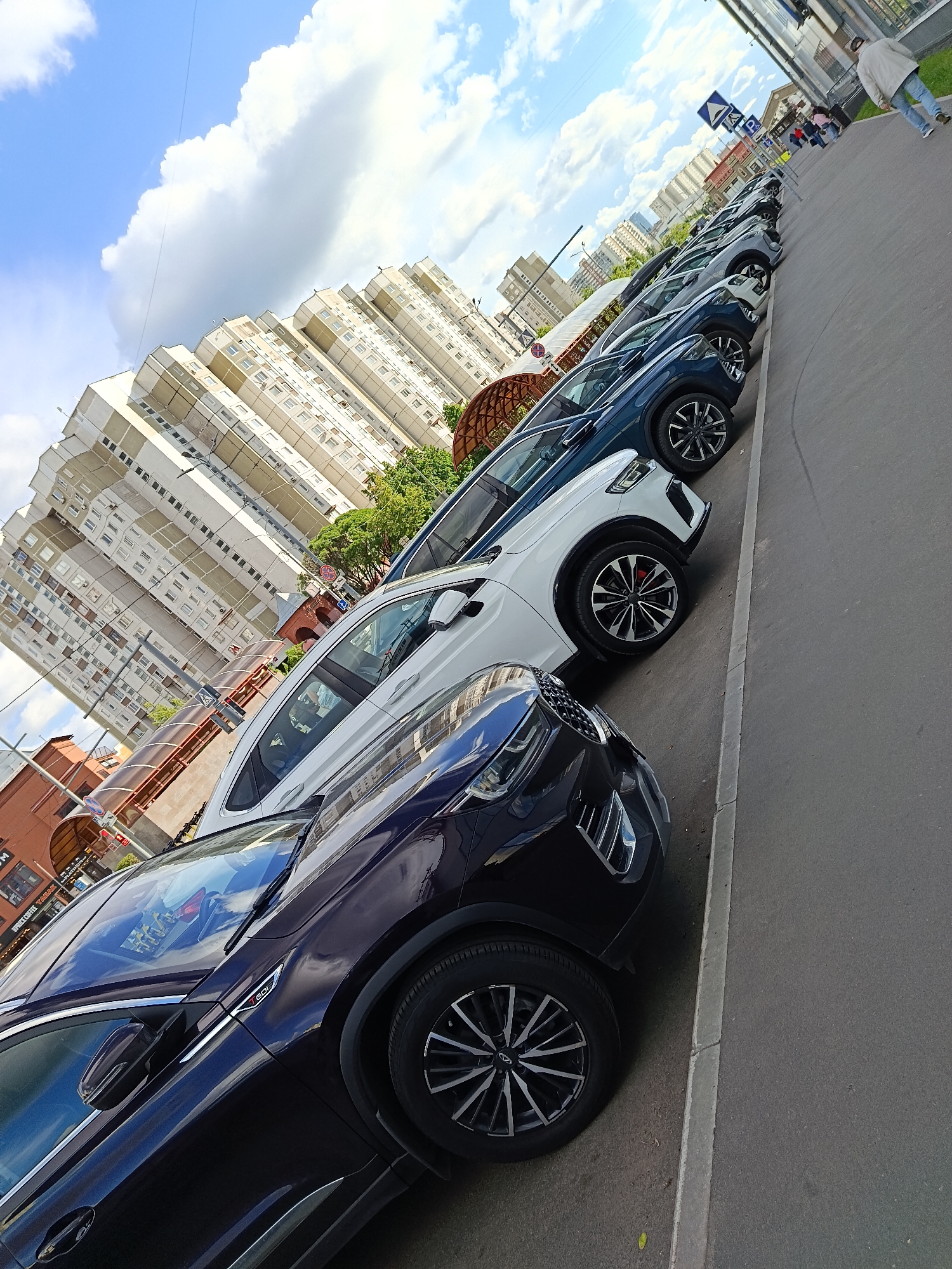 The image depicts a row of parked cars on a street in Moscow, Russia. The cars, a mix of dark blue, white, and grey, are parked parallel to the curb on the right side of the frame. A sidewalk is visible to the right of the cars. In the background, tall apartment buildings rise towards a blue sky with white clouds. A street sign is present near the end of the row of cars, with some pedestrians passing by. The scene suggests an urban environment with dense residential development.