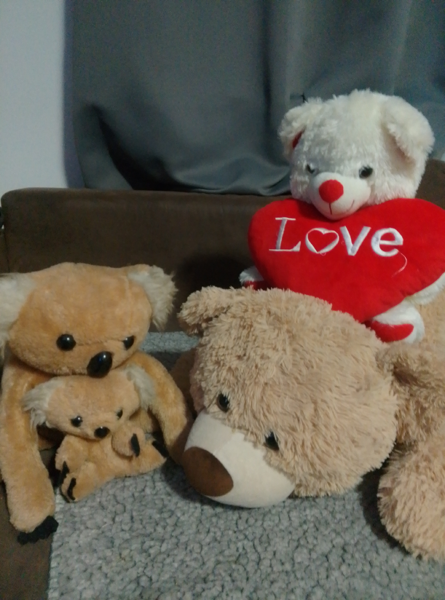 A collection of stuffed animals are arranged on a textured surface. In the foreground, a large, light brown teddy bear is lying on its side, facing right, with its head resting on the surface. To its left, a lighter brown koala bear is seated, holding a smaller, similar koala bear in its arms. Behind these, a white teddy bear with a red heart-shaped pillow that says "Love" is positioned, appearing to be seated. The background consists of a dark grey curtain or fabric and a brown upholstered surface, possibly a couch. The overall environment appears to be indoors, in a domestic setting. The location in Wedel, Germany is not discernible from the provided image.
