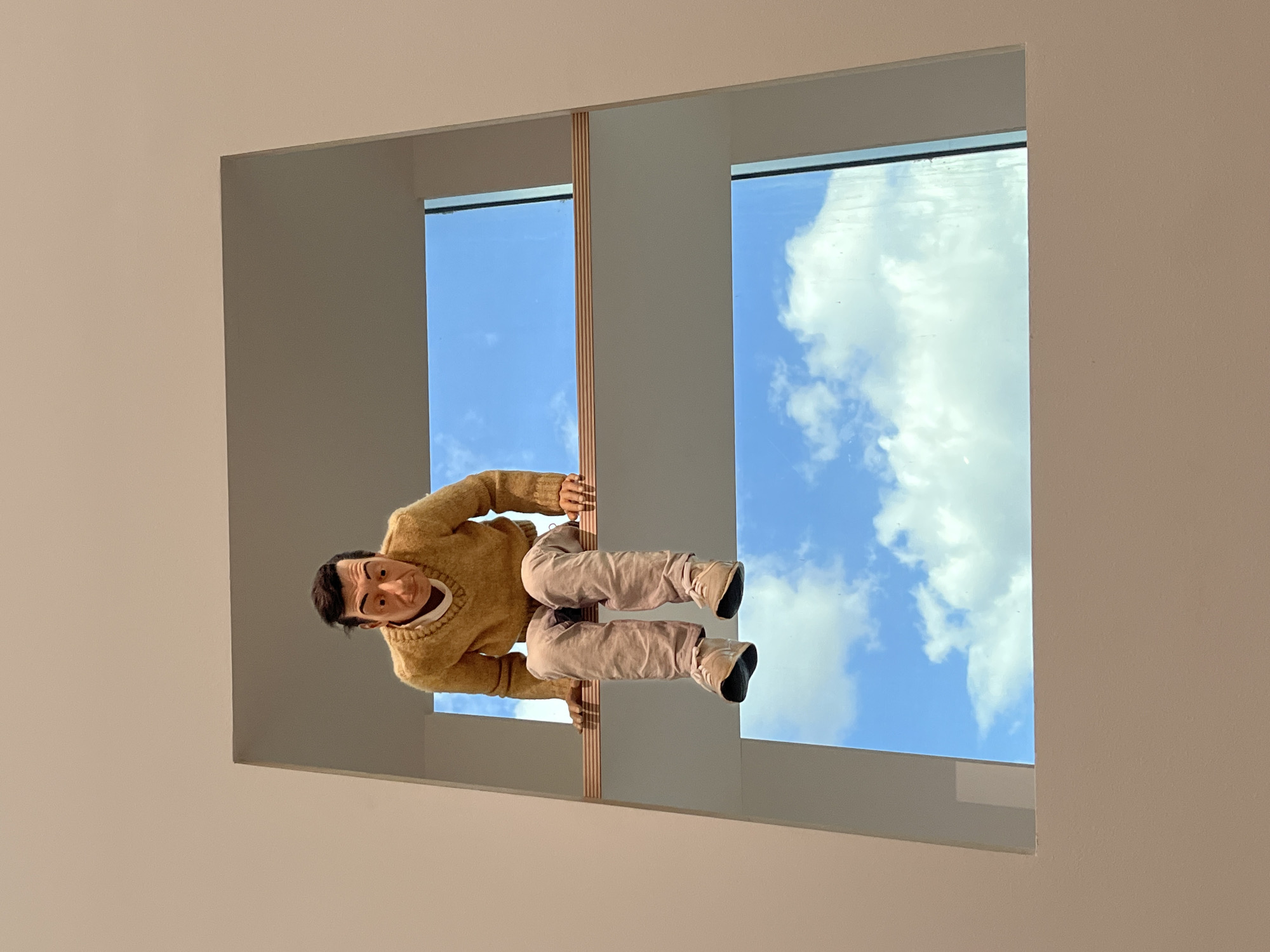 The image shows a puppet of a man wearing a tan sweater and tan pants. The puppet is suspended in the middle of a large rectangular window with a view of a blue sky with white clouds. The window is part of the Pompidou Centre-Metz, a modern art museum located in Metz, France. The window is divided into two sections by a vertical beam. The walls surrounding the window are a light beige color. The image is taken from below, looking up at the puppet and the sky.