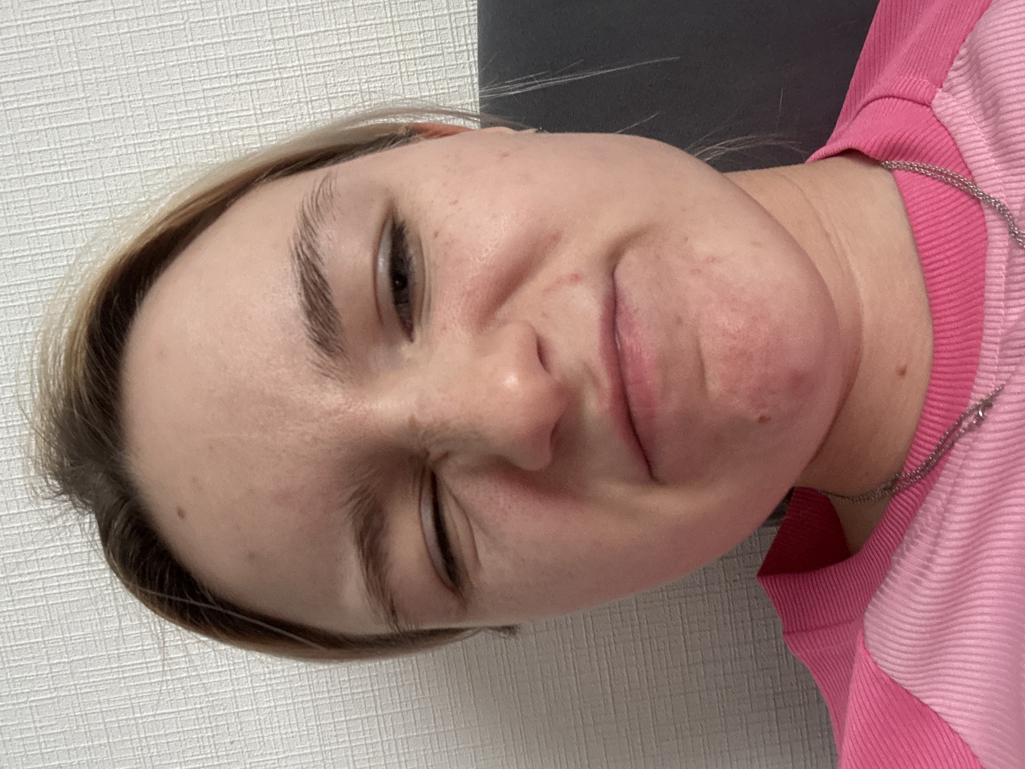A close-up portrait of a light-skinned person with light brown or blonde hair, wearing a pink and red top, and a silver necklace. The person is facing the camera at an angle with one eye closed and a slight grimace. The background consists of a white textured wall on the left and a dark or black surface on the top. Location: Unknown, Russia.