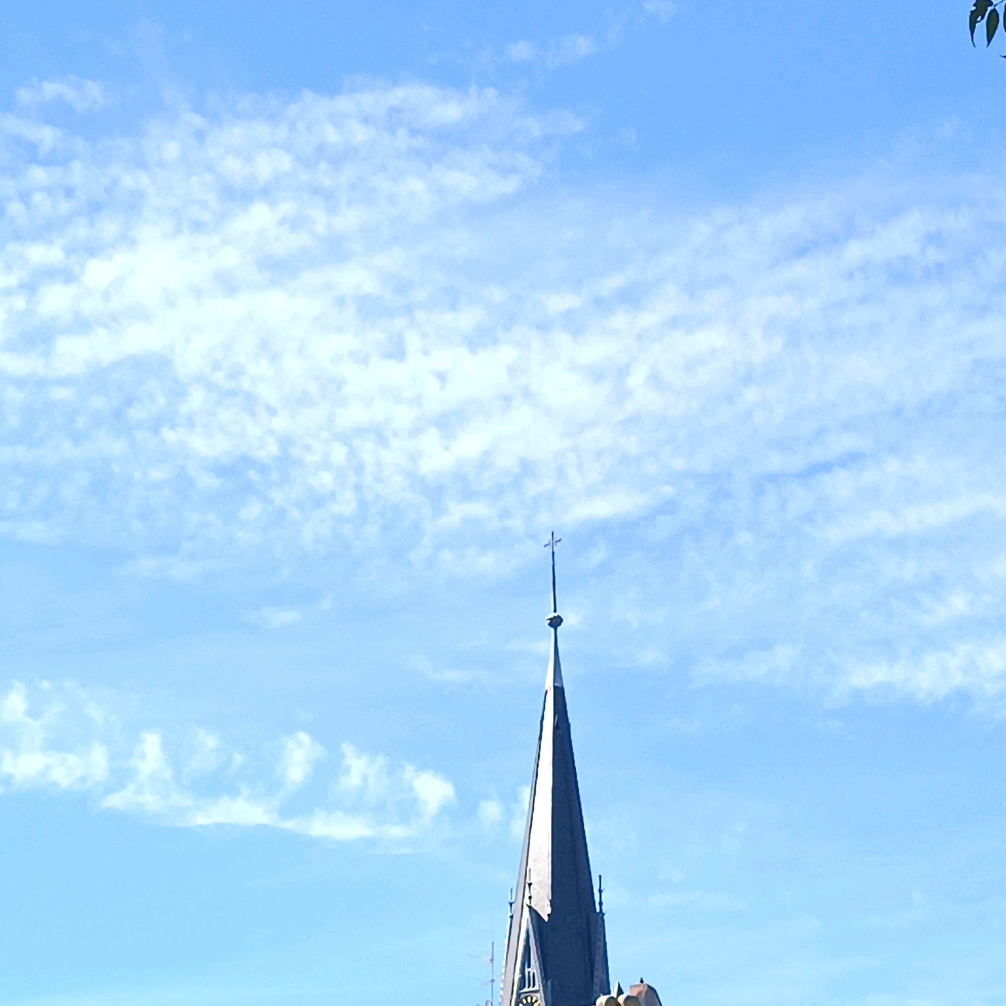 The image captures a bright, clear blue sky with scattered white, wispy clouds. Dominating the lower center of the frame is the pointed spire of a church, likely St. Mary's Church (Marktkirche) in Wiesbaden, Germany, given its distinctive architectural style. The spire is dark grey and tapers to a sharp point, topped with a small, silver cross. The spire is partially illuminated by sunlight, casting some areas in shadow. Hints of other building elements, possibly gargoyles or decorative spires, are visible at the base of the main spire. A few dark green leaves from an unseen tree are visible in the upper right corner, providing a natural element against the man-made structure and the sky. The overall scene suggests a daytime view with good weather conditions.