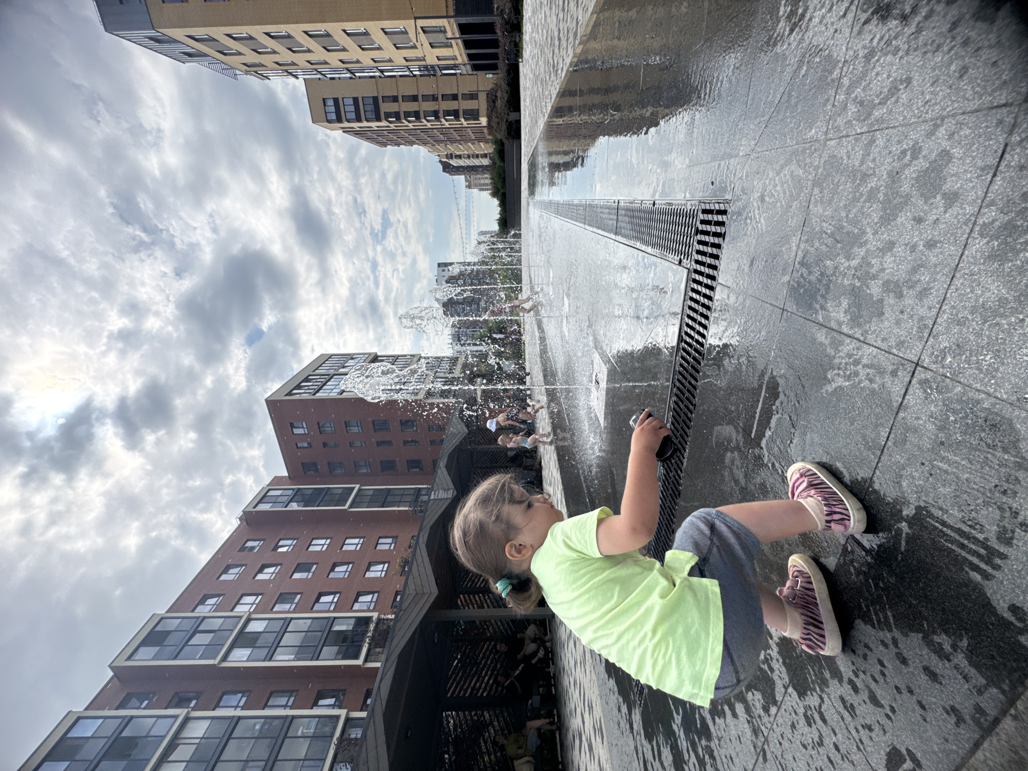 In Novoselye, Russia, a young girl in a light green t-shirt and grey shorts crouches near a public fountain with water jets spraying upwards. She wears zebra-striped pink sneakers and holds a dark object in her right hand. Other children are visible playing in the fountain in the background. The scene is set in a modern urban plaza with contemporary apartment buildings lining the sides. The ground is paved with dark tiles, wet from the fountain's spray, reflecting the cloudy sky and surrounding architecture. A covered seating area with people is visible to the left.