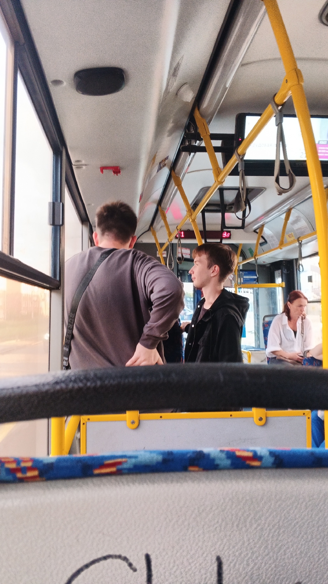 The image depicts the interior of a bus in Izhevsk, Russia. In the foreground, a black padded bar runs horizontally across the frame. Behind it, a seat with a blue and red patterned fabric is visible.
In the middle ground, two young men are standing. The man on the left is wearing a dark gray long-sleeved shirt and a black strap across his shoulder. He is facing away from the camera, towards the window. The man on the right is wearing a black hooded jacket and is looking to his left.
In the background, a woman with reddish hair is seated on the right side of the bus, wearing a white shirt. She is looking down. The bus interior features yellow grab handles and poles, with a screen displaying information visible above the yellow poles. A window on the left side of the bus allows sunlight to enter, illuminating the scene. The floor of the bus is not clearly visible, but some seating elements are present. The overall lighting suggests it is daytime, possibly late afternoon or early evening due to the warm light.