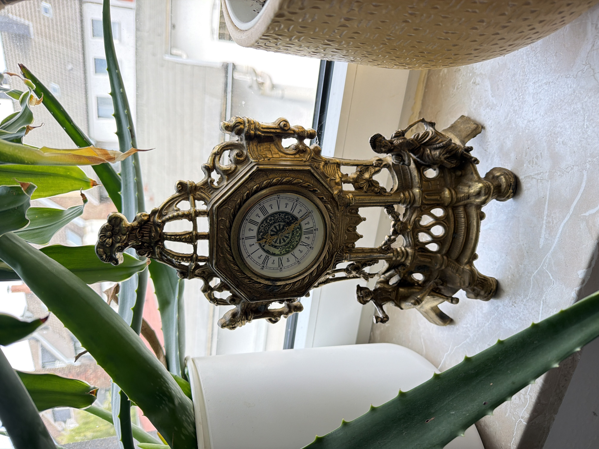 An ornate brass mantel clock with Roman numerals sits on a marble-patterned windowsill in Herne, Germany. The clock is surrounded by large, green aloe vera plants in white and tan pots, with a blurred view of houses and a cloudy sky visible through a window in the background.