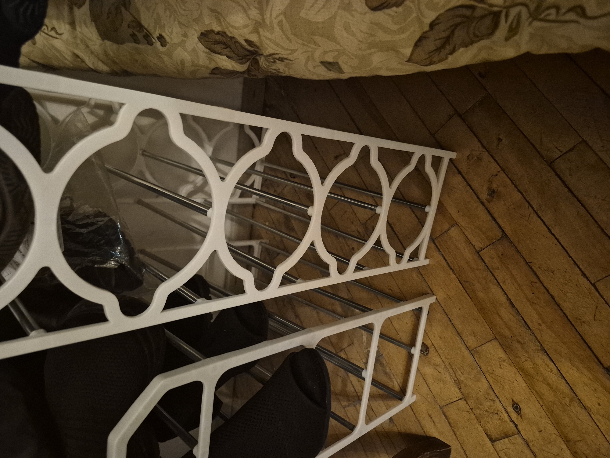 The image presents an indoor scene focused on a white modular shoe rack with two visible tiers. The rack is constructed from white plastic or similar material, featuring decorative, interconnected circular patterns on its side panels, and holds shoes on silver-colored metal rods. Several dark-colored shoes or slippers are stored on and below the rack, including what appears to be black knitted slippers and other shoes partially covered in clear plastic.
Above the shoe rack, the edge of a bed or furniture piece is visible, adorned with a beige and brown patterned fabric, possibly a bedspread or blanket with leaf motifs. The floor, visible on the right side of the frame, is made of aged wooden planks with prominent grain patterns, knots, and signs of wear, running diagonally. The lighting is dim, characteristic of an indoor setting. No people are present, and no specific activity is occurring. There is no legible text visible on any of the objects. While the location context is given as Yerevan, Armenia, the image itself does not contain any specific visual cues to confirm this location.