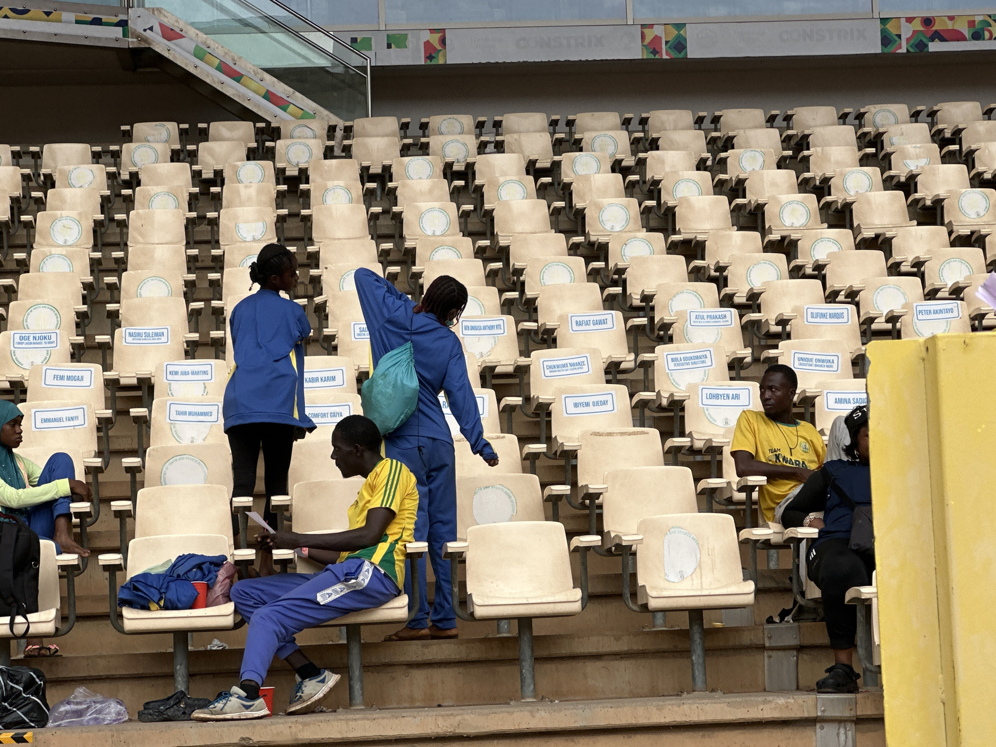 The image shows a partially filled seating section in an outdoor stadium. Several individuals are visible in the foreground: two wear matching blue tracksuits, one holding a turquoise bag; another wears a yellow and green shirt with blue track pants and sits reading a paper. To the left, a woman in a light green top and blue pants sits with a backpack. To the right, a person in a yellow shirt sits with a woman in a dark top and black pants. The seats are beige with white placards, each bearing names such as "OGE NJOKU," "NASIRU SULEIMAN," "RAFIAT GAWAT," and "CHUKWUMA NWANZE." The placards also display the logo of the "Ministry of Youth and Sports Development." The background reveals rows of empty seats, leading to the upper section of the stadium with a sign that reads "CONSTRIX".