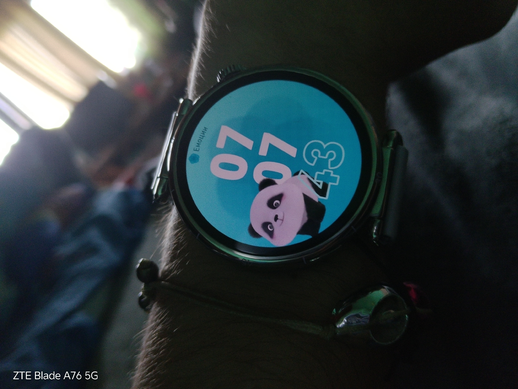 The image shows a close-up of a person's wrist wearing a smartwatch. The smartwatch has a bright blue circular display with a cartoon panda character at the bottom. Large pink numbers, "07" and "07," are displayed prominently on the screen, along with a "+3" to the right. A small word, "Емоции," which translates to "Emotions" in Russian, is visible on the left side of the screen. A silver bracelet with a small, metallic charm is also visible on the wrist, below the watch. The background is dark and out of focus, with some bright light sources suggesting an indoor environment. The text "ZTE Blade A76 5G" is overlaid at the bottom left corner of the image. The location is not discernible from the image.