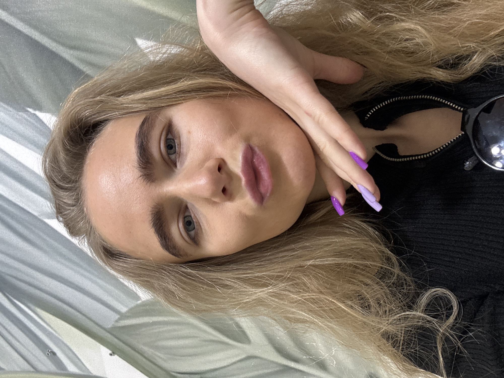 A portrait of a fair-skinned female with long, light blonde hair is presented. Her left hand rests on her cheek, partially obscuring her face. She has light blue eyes, prominent eyebrows, and full lips. Her nails are painted in a lavender gradient. She wears a black top with a visible zipper and part of a pair of sunglasses can be seen near her collar. The background is a patterned surface. The location is Unknown, Unknown.