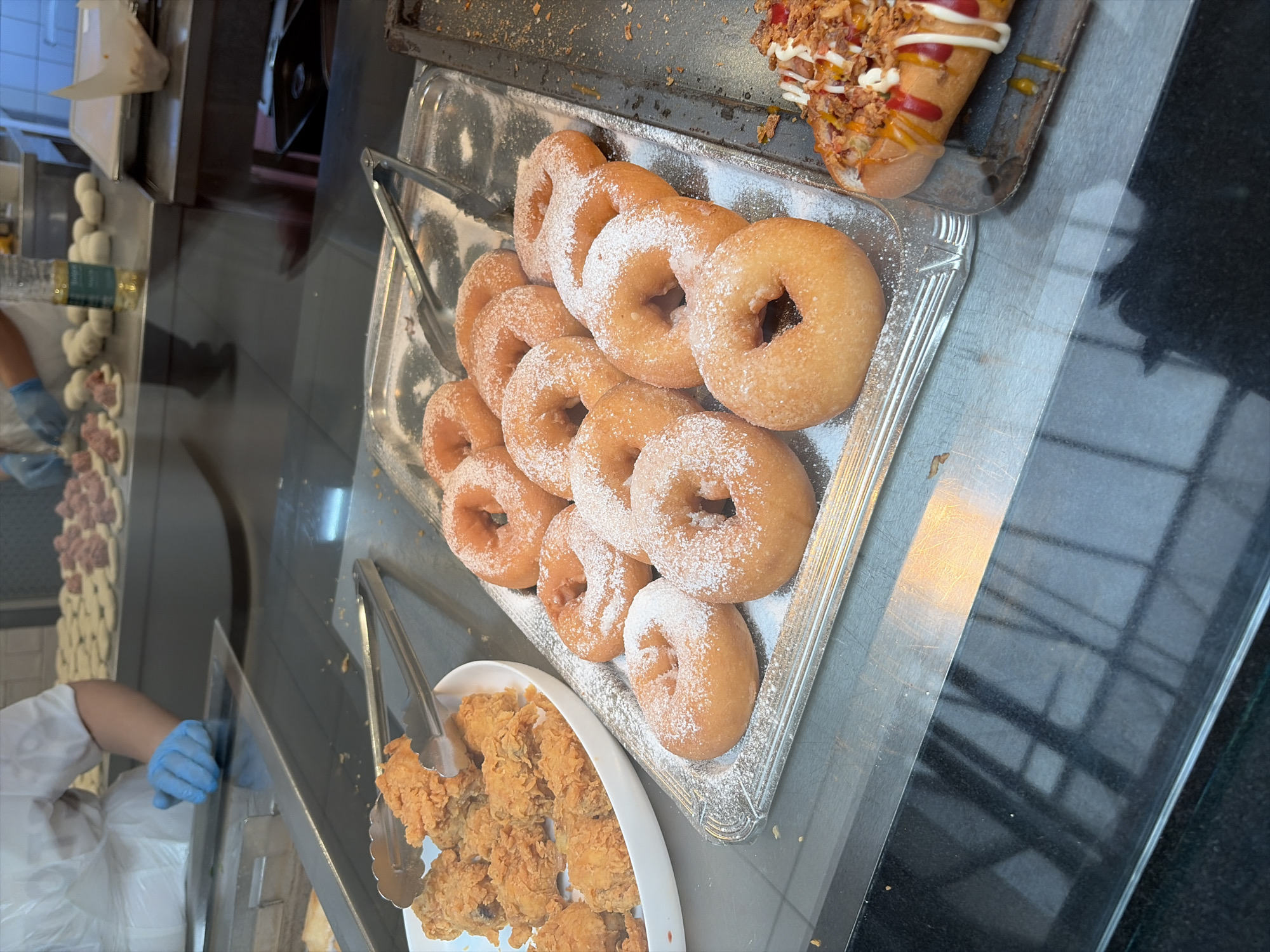 In Balashikha, Russia, a food service area features a display of fried chicken and donuts. A person in a white chef's coat and blue gloves is partially visible in the background, arranging food items on a stainless steel counter. The foreground shows a white plate of crispy fried chicken next to a rectangular silver tray filled with powdered sugar-dusted donuts. To the right, another tray holds a hot dog topped with condiments and crispy onions. The overall scene suggests a self-serve food establishment or buffet.