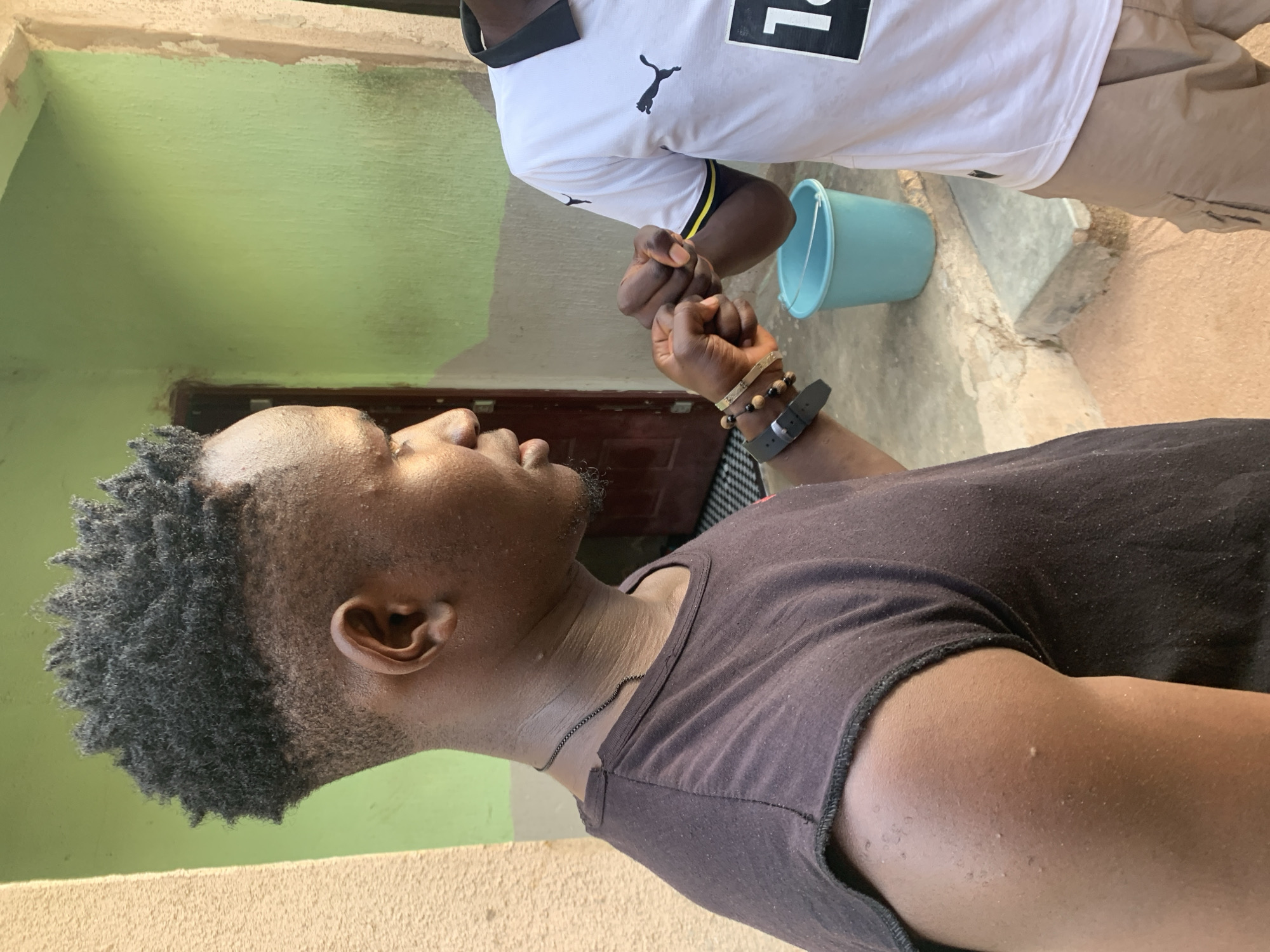 The image shows two individuals in what appears to be an outdoor or semi-outdoor setting in Jalingo, Nigeria. One person is wearing a black t-shirt and a thin chain necklace. They have short, dark, curly hair and a goatee. They are wearing a watch and bracelets on their left wrist. The other person is wearing a white Puma t-shirt with black and yellow trim on the collar. They are wearing light-colored pants. Both individuals are shown from the chest up.
The two people are performing a fist bump. Behind them, a light blue bucket is visible. The background includes a light green wall with some peeling paint at the top, and a darker brown door. The ground appears to be concrete or paved. The lighting suggests it is daytime. The overall mood appears casual and friendly, as indicated by the gesture of a fist bump. There is no visible text other than the Puma logo on the t-shirt.