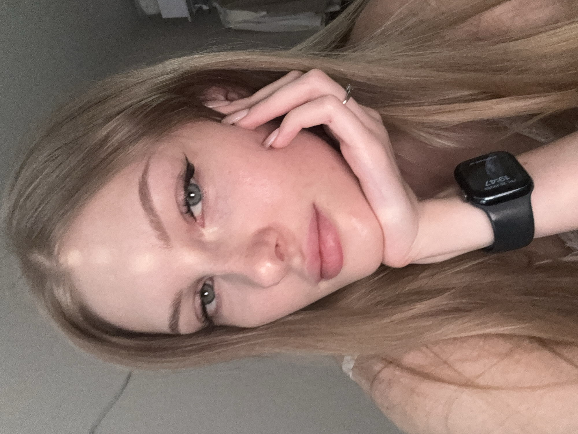 A close-up, waist-up selfie of a young Caucasian woman with long, blonde hair is shown. She is facing the camera, with her right hand resting on her chin and her left hand touching her face. She is wearing a black smartwatch on her left wrist. The background is blurred and appears to be an indoor setting, possibly a room with clothing on hangers visible on the right side. The lighting suggests it is indoors. There is no visible information to suggest this image is located in the city of Perm, Russia.