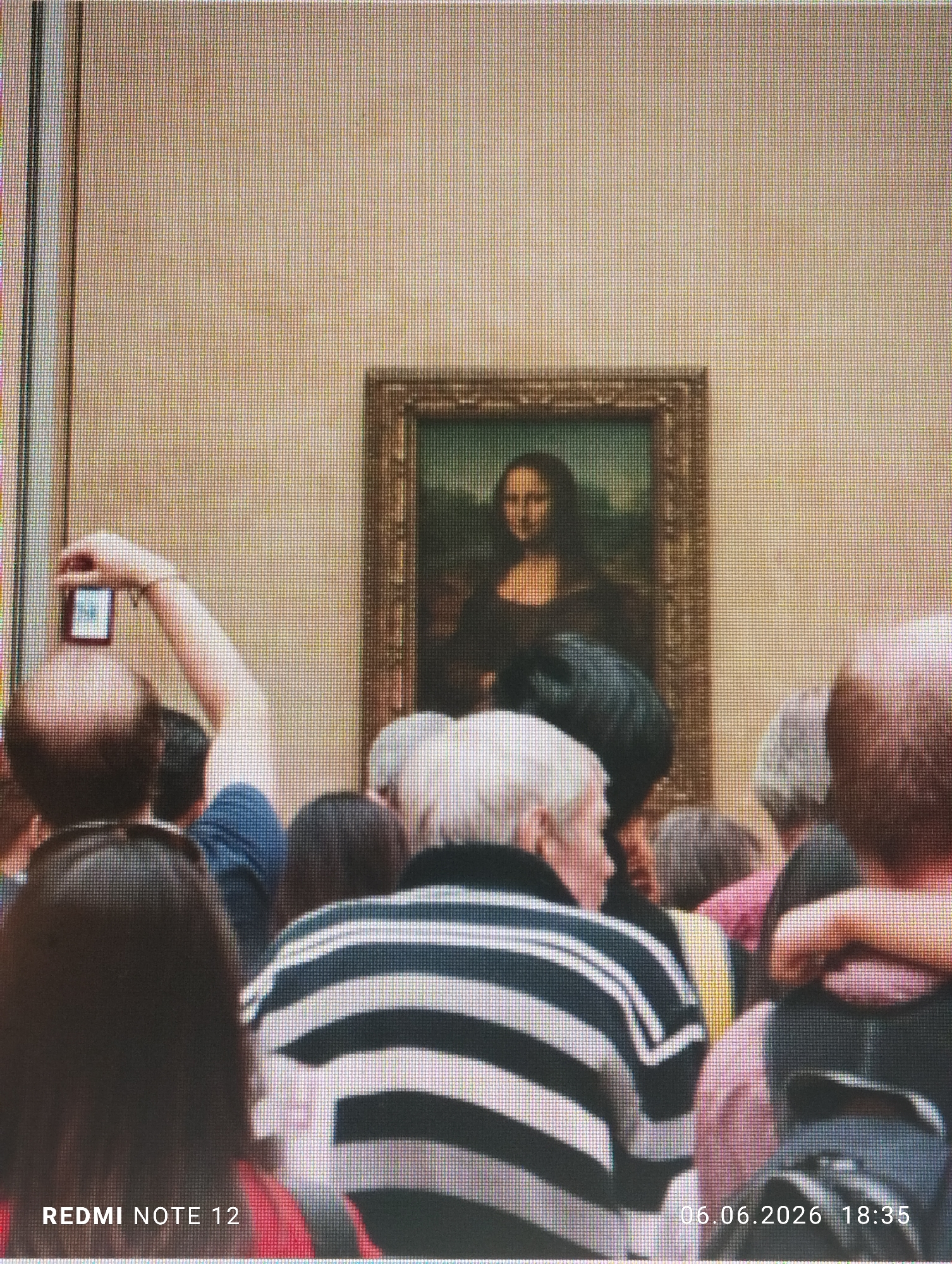 This image depicts a crowded indoor scene, identified as being within the city of Burgas, Bulgaria. The photograph itself exhibits a prominent pixel grid across its entirety, suggesting it is a capture of a digital display rather than a direct photograph of the physical environment.
The central focus of the displayed scene is a reproduction of the Mona Lisa painting, prominently positioned in the upper half of the frame. The painting is encased in a dark, ornate frame and is mounted against a plain, light beige wall with a subtle texture.
In the foreground and midground, numerous individuals are gathered, primarily viewed from behind, all facing the painting. On the left side of the frame, a person with short, dark hair extends their right arm upwards, holding a smartphone to photograph the Mona Lisa. The phone's screen clearly shows a miniature rendition of the painting. To the right, another individual wears a black and white horizontally striped polo shirt, also facing the painting. Other discernible individuals exhibit various hair colors, including grey, and wear clothing in shades of blue, pink, and red. The density of the crowd indicates a popular viewing event.
Two text overlays are present: "REDMI NOTE 12" is visible in the bottom-left corner, and "06.06.2026 18:35" is in the bottom-right corner.