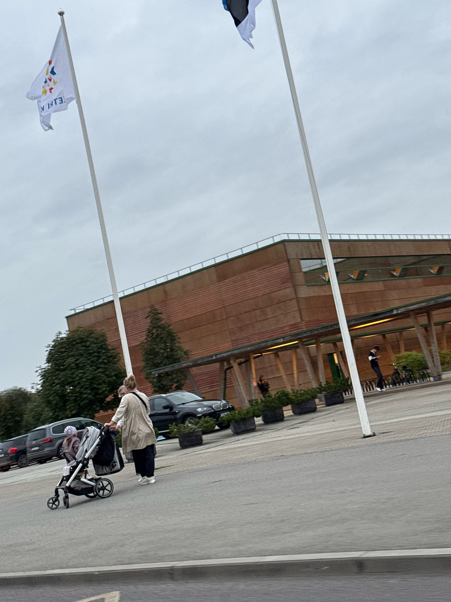 In the city of Peetri alevik, Estonia, on an overcast day, a woman pushes a stroller containing a young child. They are walking on a paved area in front of a large, modern building with a reddish-brown facade and a covered walkway. Several parked cars are visible, including a black SUV. Two flags are flying from tall poles; one is white with a design and text, and the other is partially visible and appears to be the Estonian flag. The environment suggests an outdoor public space, possibly near a cultural or governmental institution.