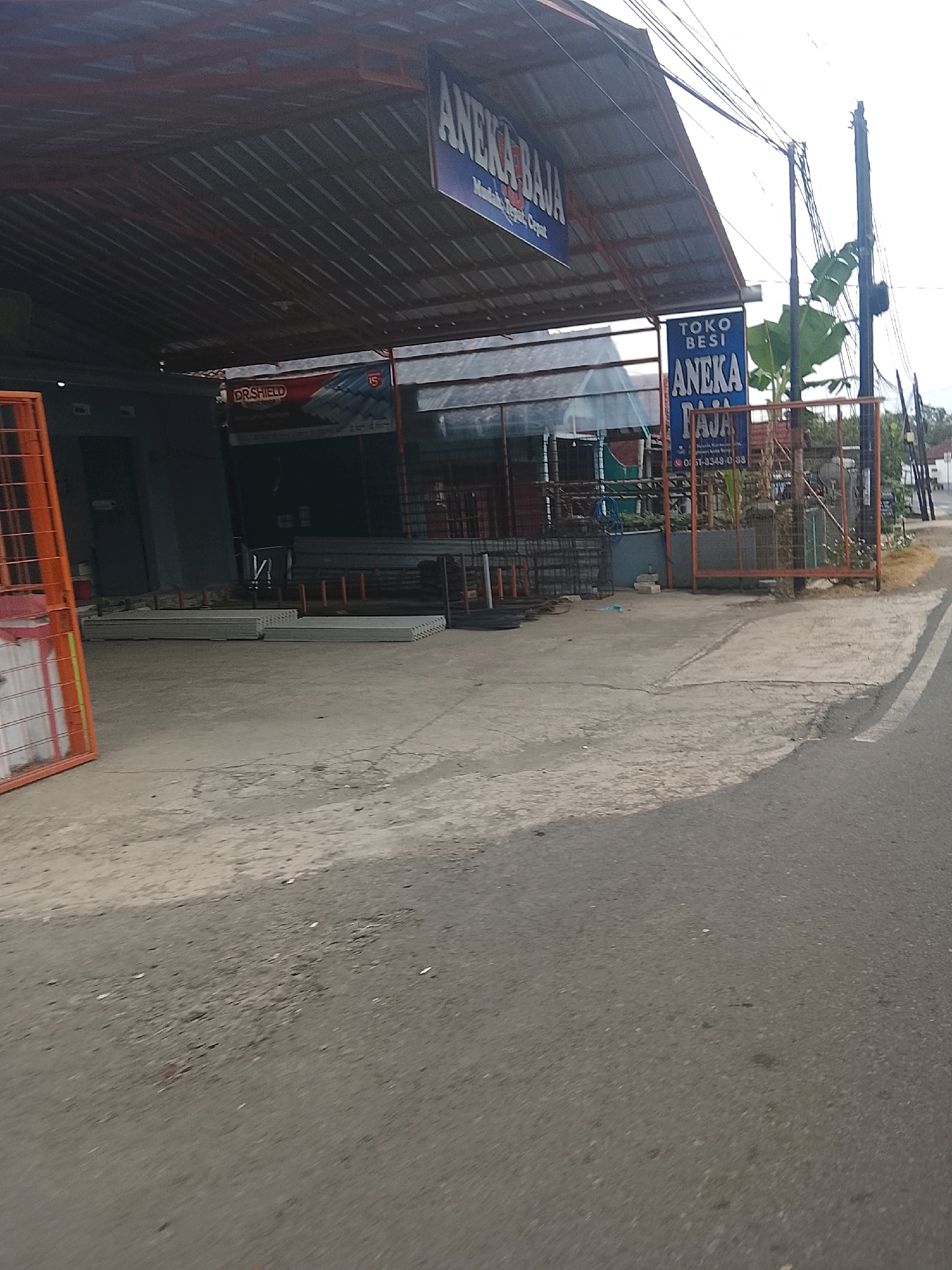 The image shows a street view of a building with a metal roof, identified by signage as "ANEKA BAJA". The building appears to be a hardware or building supply store, with various construction materials visible in front and inside. Specifically, there are stacks of what appear to be concrete panels or slabs and some metal rebar. A red metal fence is also present in the foreground, along with an orange safety barrier. Power lines are strung overhead, and a banana tree is visible to the right of the building. The street in front of the building is paved and shows some wear and tear. The overall impression is that of a commercial establishment on a street in Indonesia, specifically Banjar, given the text on the signage.