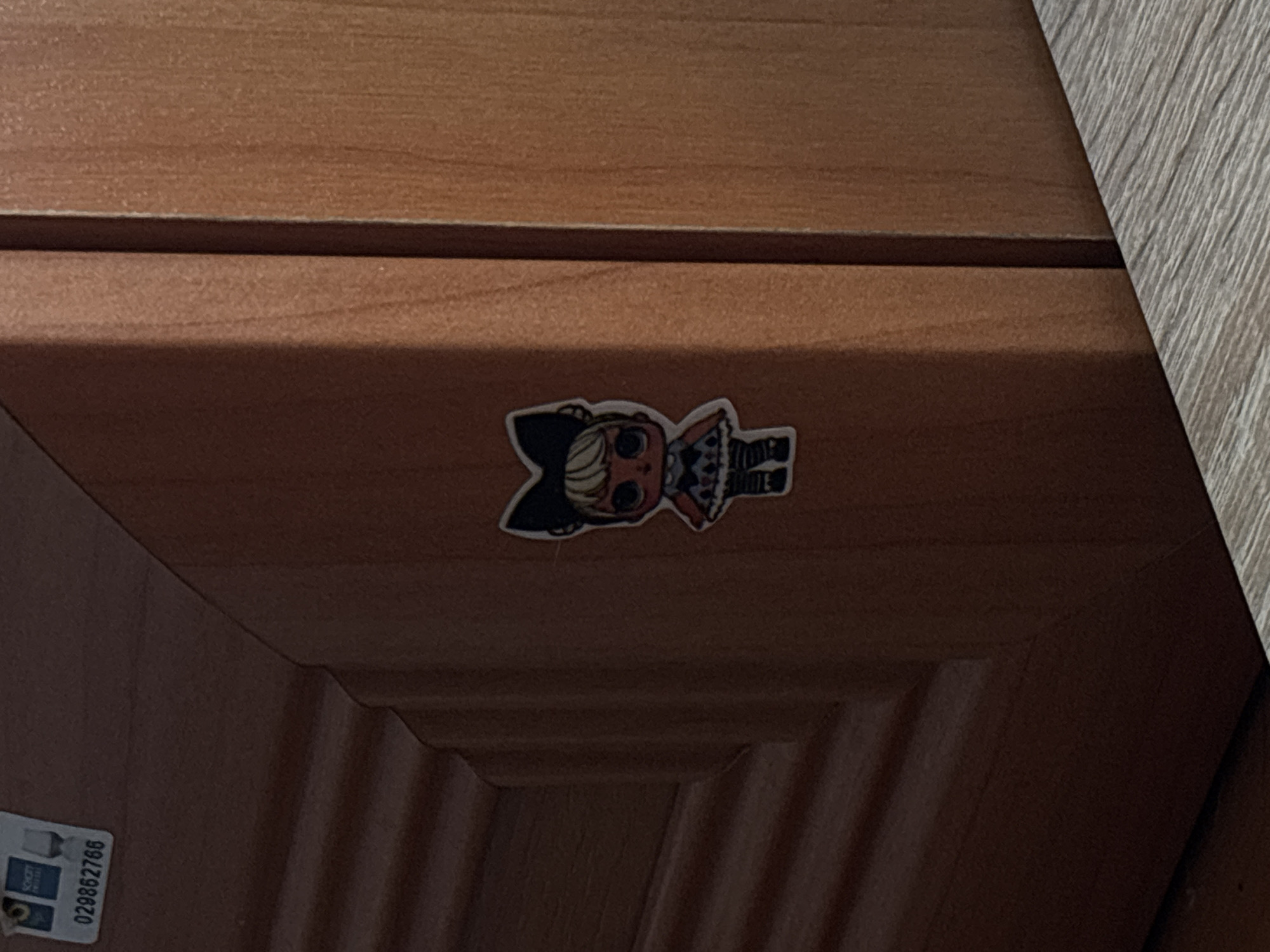 The image shows a close-up of a wooden surface, likely a piece of furniture like a drawer or cabinet, with a sticker of a stylized doll placed on it. The doll has large eyes, black hair styled with cat-like ears, and is wearing a patterned dress and boots. The sticker is colorful, with shades of orange, black, white, and blue visible. The wooden surface has a visible grain pattern and appears to be a warm, medium brown color. There is a slight shadow cast on the lower part of the surface, suggesting the lighting source is from above and slightly to the right. In the lower left corner of the image, a small white label with some text and a graphic is partially visible. The text "029862766" is clearly legible on this label, oriented vertically. The background is a lighter, textured material, possibly a wall or another piece of furniture, with vertical lines. The scene is static, with no discernible activity taking place. The context provided places this image in Saint Petersburg, Russia, though there are no specific visual cues within the image itself to confirm this location.