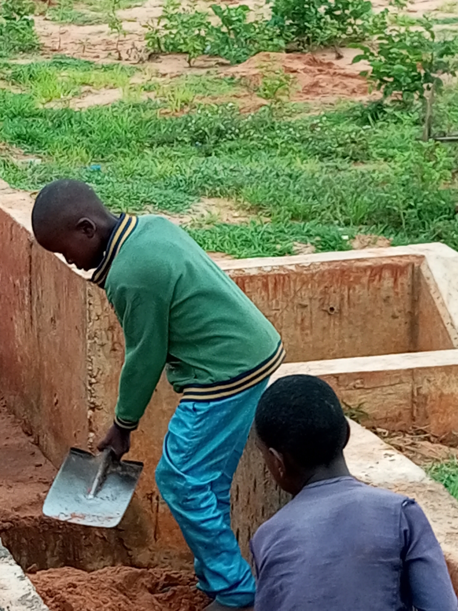 The image depicts two young boys near a construction site in Gombe, Nigeria. The boy in the foreground, on the left, is wearing a green jacket with yellow and blue trim and blue pants. He is holding a shovel and appears to be engaged in digging or moving earth. The second boy, on the right, is wearing a dark purple long-sleeved shirt and is facing away from the camera. They are positioned next to a concrete structure with reddish-brown walls, possibly a foundation or a water reservoir. The surrounding environment consists of dry, sandy soil and sparse green vegetation, indicative of an outdoor setting in a dry climate. The overall scene suggests an activity related to construction or land preparation.