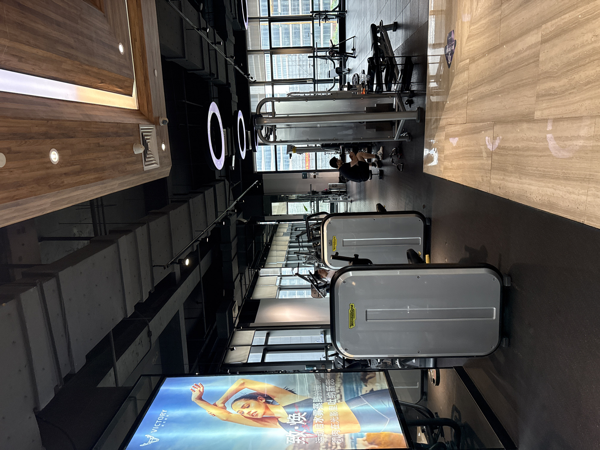 The image depicts the interior of a modern gym located in Haizhu District, China. The gym features a variety of exercise equipment, including weight machines, benches, and free weights, arranged on a dark rubberized floor. Large windows line one side of the gym, offering a view of urban buildings. A large digital display board on the left side of the image shows an advertisement for "VICTORY." Two individuals are present in the gym: one person is seated on a weight machine, appearing to be resting or preparing for an exercise, while another person is standing near a different machine, possibly engaged in a workout. The lighting is a mix of overhead track lighting and decorative circular light fixtures. The ceiling is a combination of exposed industrial elements and decorative wooden panels. The floor near the foreground is tiled with a light-colored, wood-grain pattern.