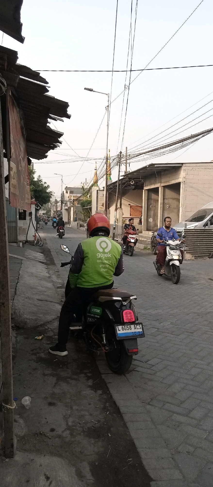 A rear view shows a person wearing a red helmet and a green Gojek jacket, sitting on a black motorcycle. The motorcycle has a license plate reading "L 6058 ACS". The rider is facing away from the camera, looking down a street. Several other motorcycles and riders are visible further down the street, some heading in the same direction. On the right side of the street, there is a white van and a building with a corrugated metal roof. The street is paved with interlocking bricks. In the background, two minarets of a mosque are visible, and power lines crisscross the sky. The scene appears to be in Wadungasri, Indonesia, suggested by the style of buildings and the presence of a Gojek rider.