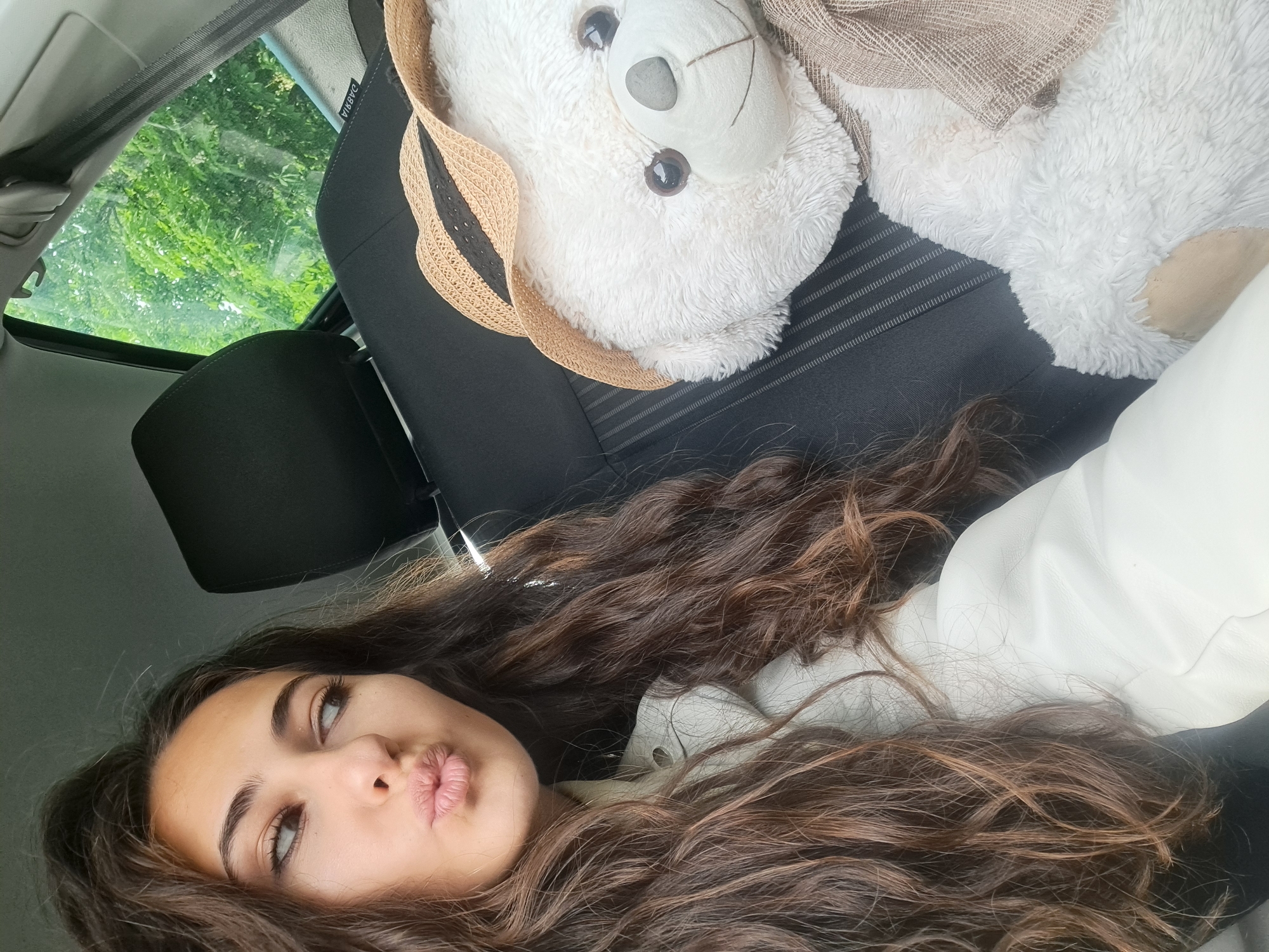 A selfie shows a young woman with long, wavy brown hair and pursed lips lying in a car, next to a stuffed white bear wearing a straw hat and scarf. The car seat is black with vertical stripes. A window showing green trees is visible in the background. The location is not specified.