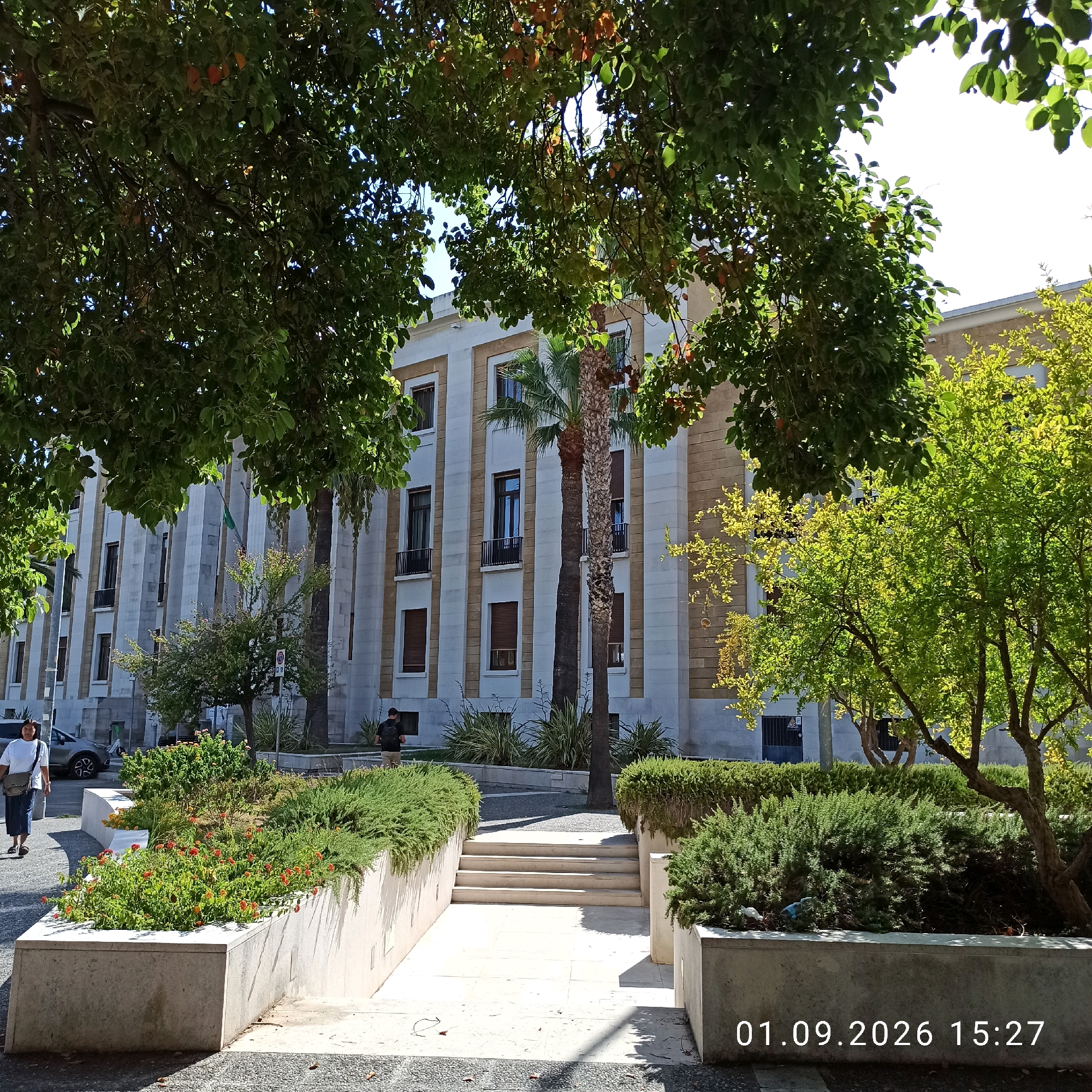 The image shows a street scene in Bari, Italy, in front of a large, modern building with a beige and white facade. The building has multiple floors with numerous windows, some of which are covered by shutters. Several trees with green foliage are visible, with one prominent palm tree located in front of the building. In the foreground, there is a landscaped area with plants and flowers, bordered by concrete planters. A set of stairs leads down into a paved area. Two individuals are present: one person, appearing to be a woman, walks from the left side of the frame towards the foreground, carrying a bag over her shoulder. Another person, a man, is seen walking away from the camera in the middle ground. The ground is paved with a combination of asphalt and cobblestones. The date and time "01.09.2026 15:27" are overlaid on the lower right corner of the image.