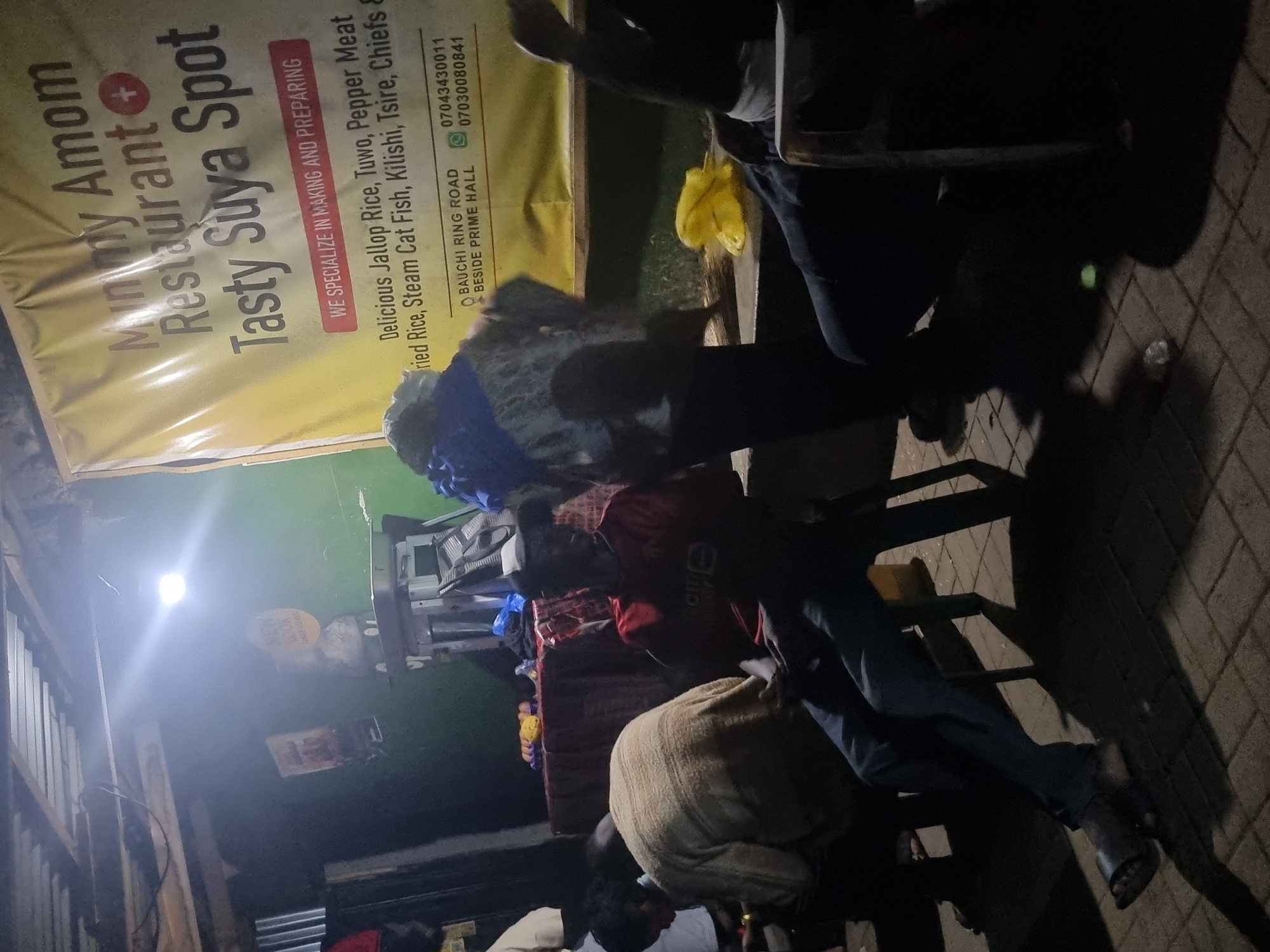 In Gwong, Nigeria, a dimly lit scene unfolds at an outdoor restaurant. A large yellow banner with red text dominates the background, advertising "Mummy Amom Restaurant Tasty Suya Spot." The banner lists menu items including "Delicious Jallop Rice, Tuwo, Pepper Meat, Fried Rice, Steam Cat Fish, Kilishi, Tsire, Chiefs &." Below this, contact information and a location are provided: "BAUCHI RING ROAD BESIDE PRIME HALL" and phone numbers "07043430011" and "07030080841."
Several people are present in the foreground, with varying degrees of visibility. A man in a red t-shirt and blue jeans is seated on a chair, facing to his left, with his legs crossed. To his left, another person in a light-colored sweater is seated, their back mostly to the camera. In the shadows on the far right, another individual is partially visible, seated and leaning forward. The ground is paved with interlocking bricks, and shadows are cast by the overhead lighting and the structures around the establishment. The overall atmosphere is casual and appears to be a gathering at a local eatery.